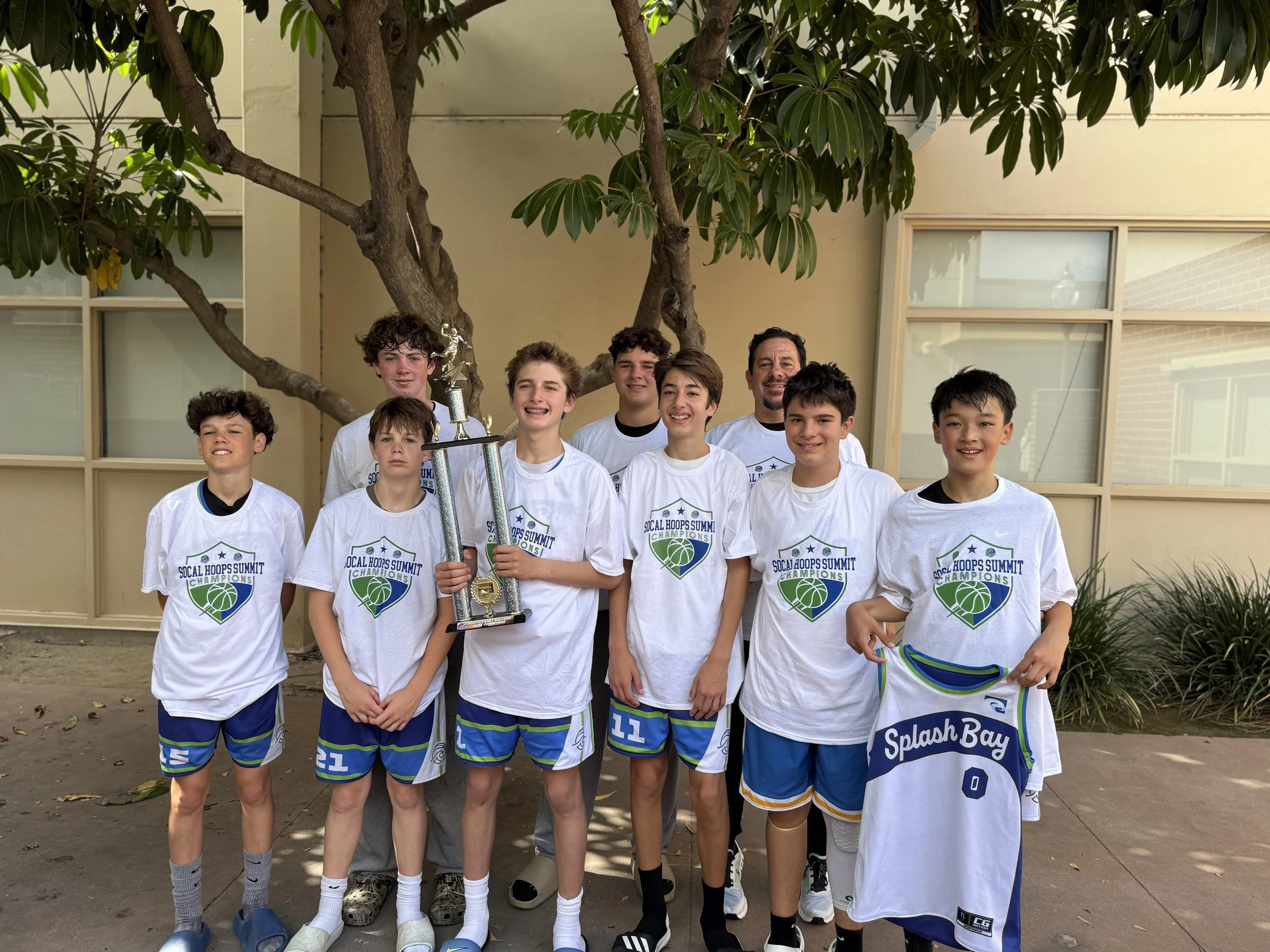 A youth basketball team standing outdoors under a tree, holding a large trophy and a team jersey, celebrating their victories. They are wearing white team shirts with a basketball logo and the words 'Soccal Hoops Summit Champions'.