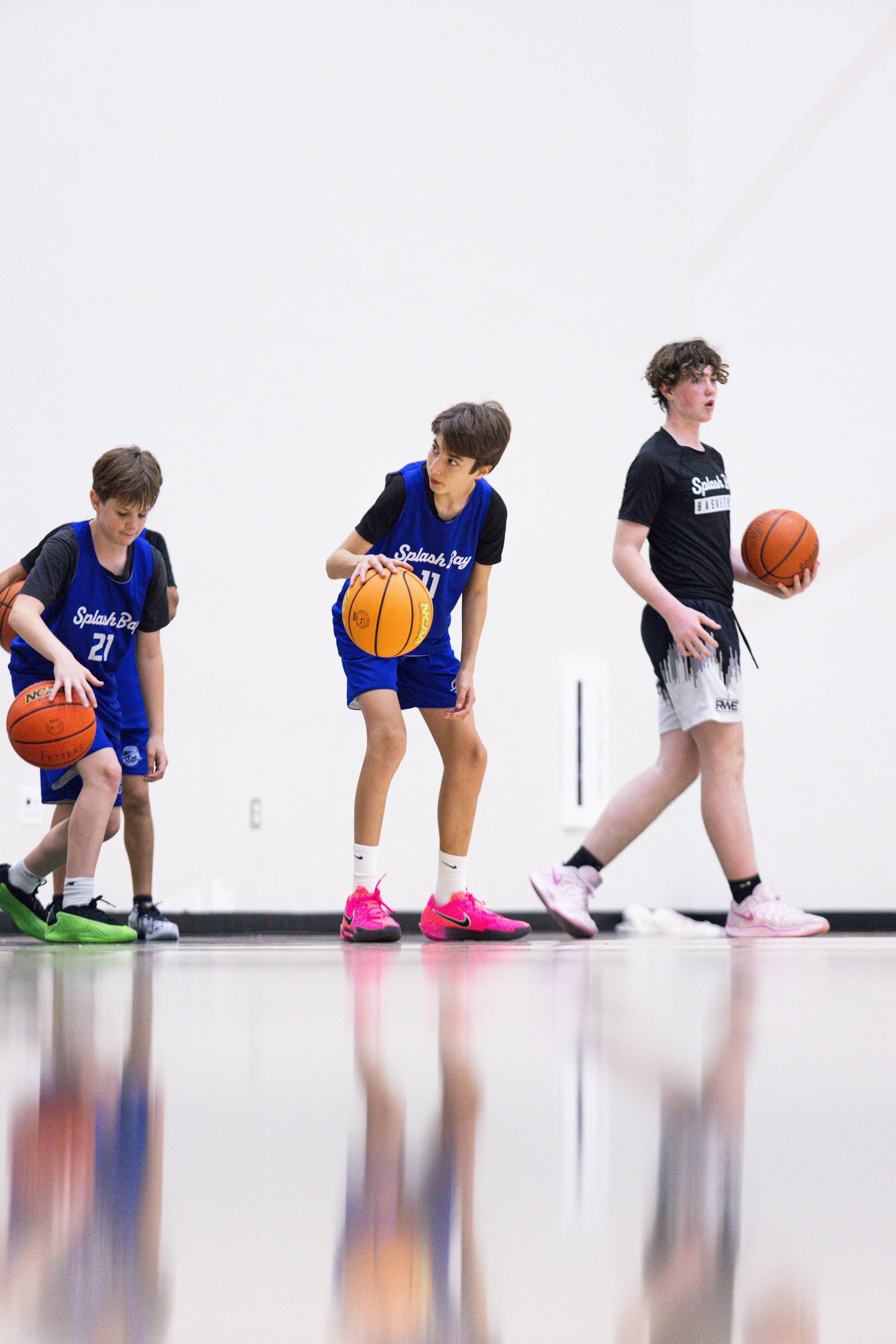 Three boys practicing basketball on an indoor court, standing in a line, with the middle boy in a blue jersey and pink shoes, holding a basketball.