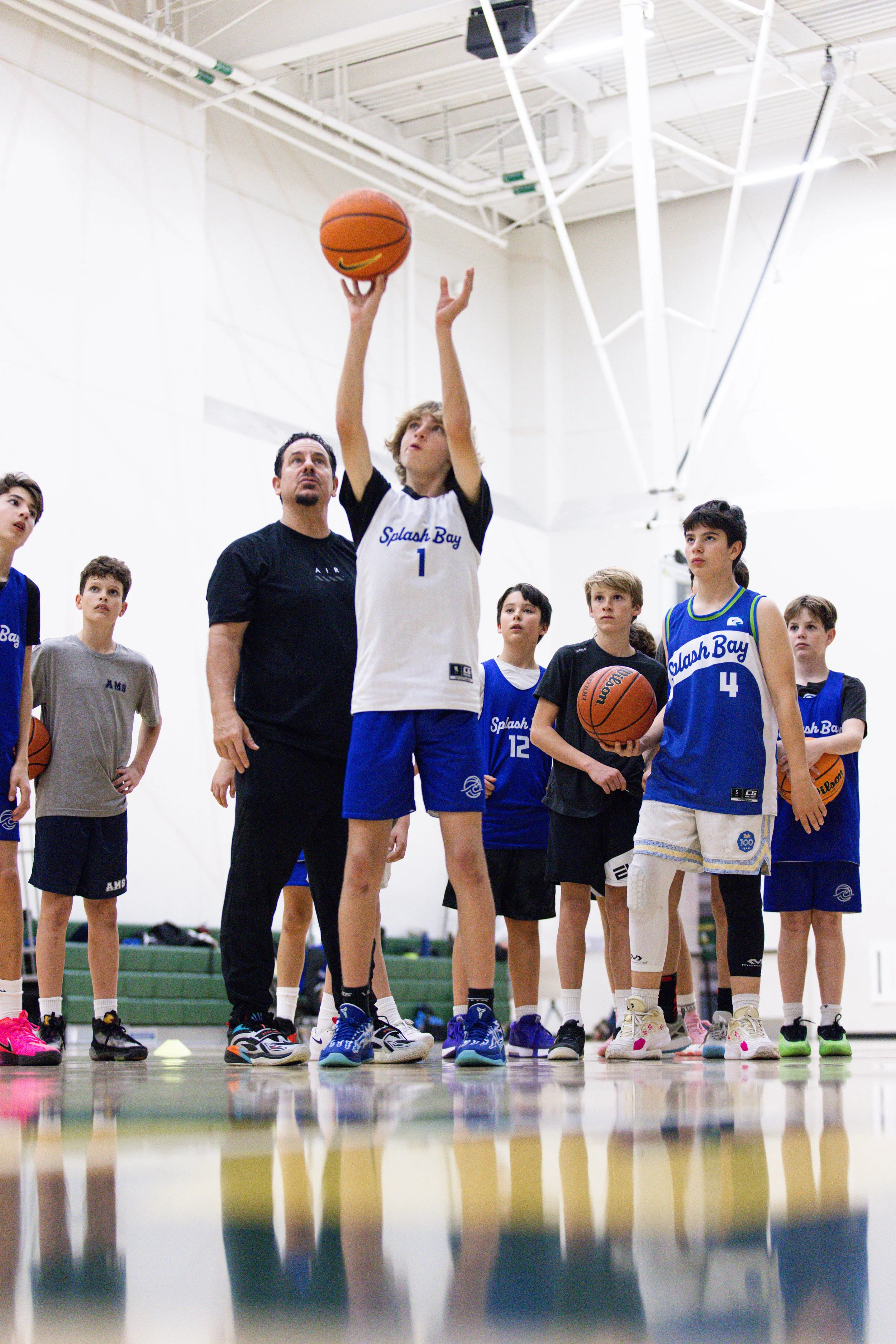 Young basketball player in white jersey taking a shot in gymnasium surrounded by children and coach.