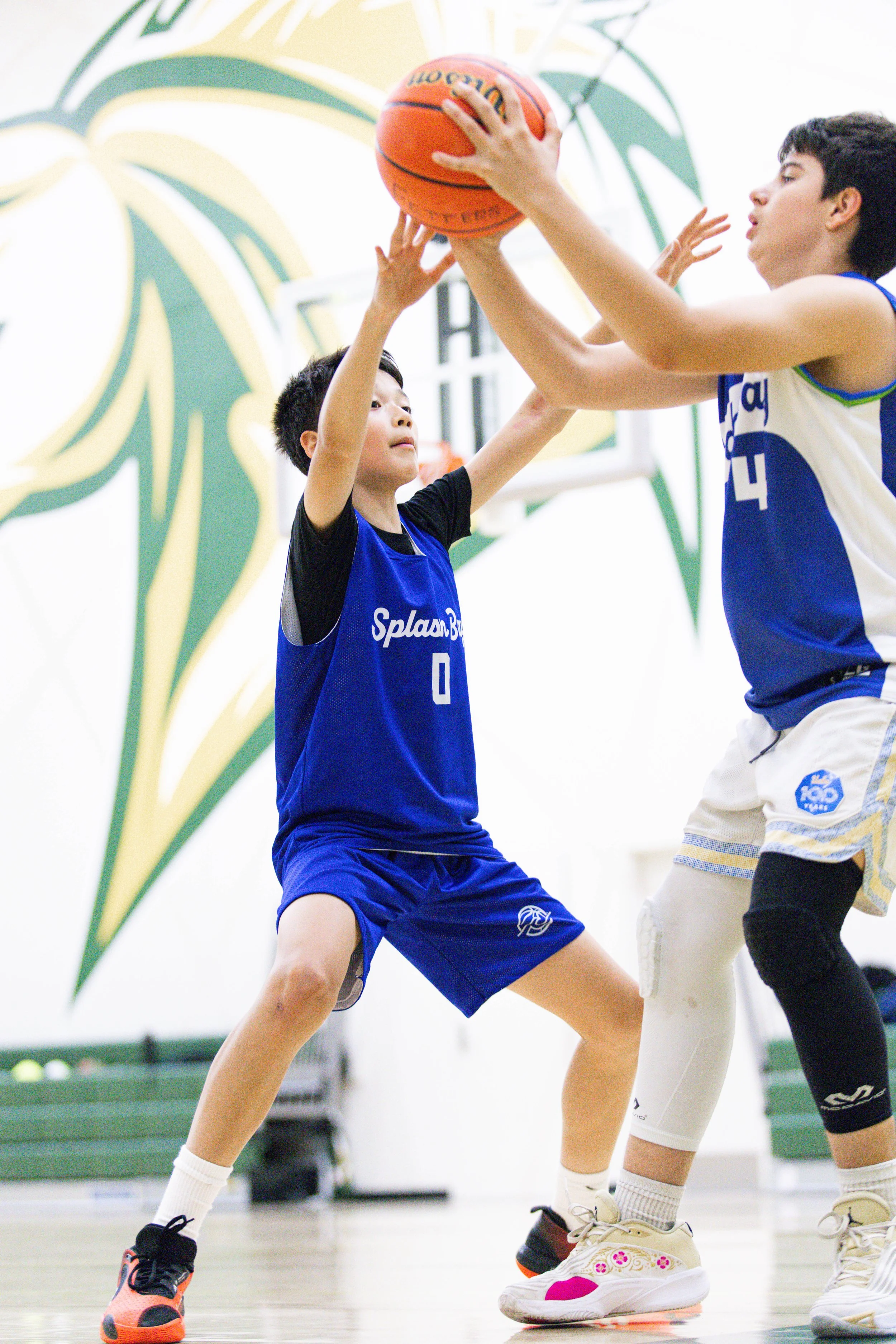 Two young boys playing basketball indoors. One boy in a blue jersey is holding the ball and attempting to block the other boy's shot. The background features a colorful mural on the wall.