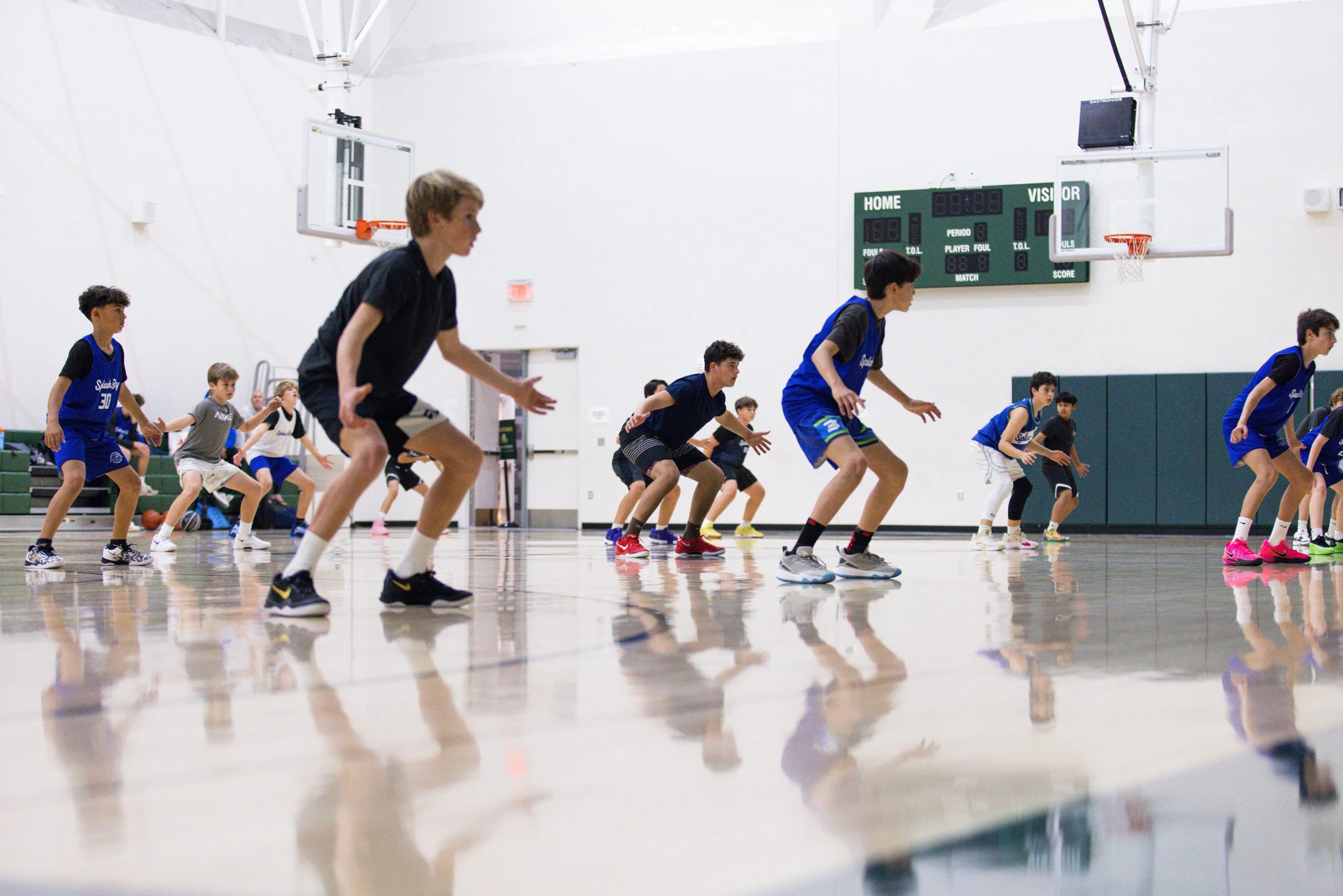 A group of young boys in athletic clothing practicing basketball drills on an indoor court, with some boys crouching and others standing in a staggered formation, reflected on the polished wooden floor.