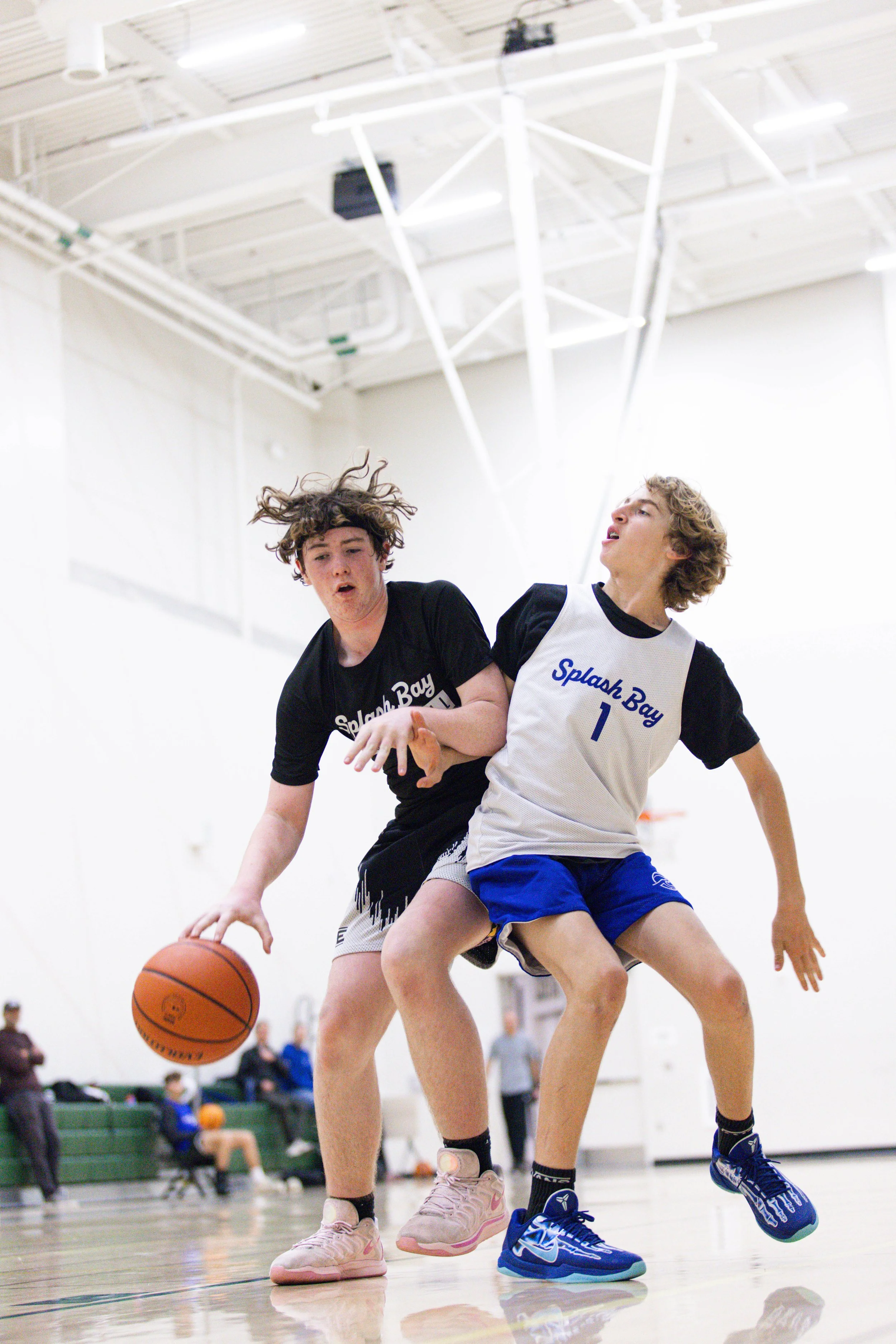 Two boys playing basketball indoors, one wearing black shirt and the other in a white and black jersey numbered 1, competing for the ball.