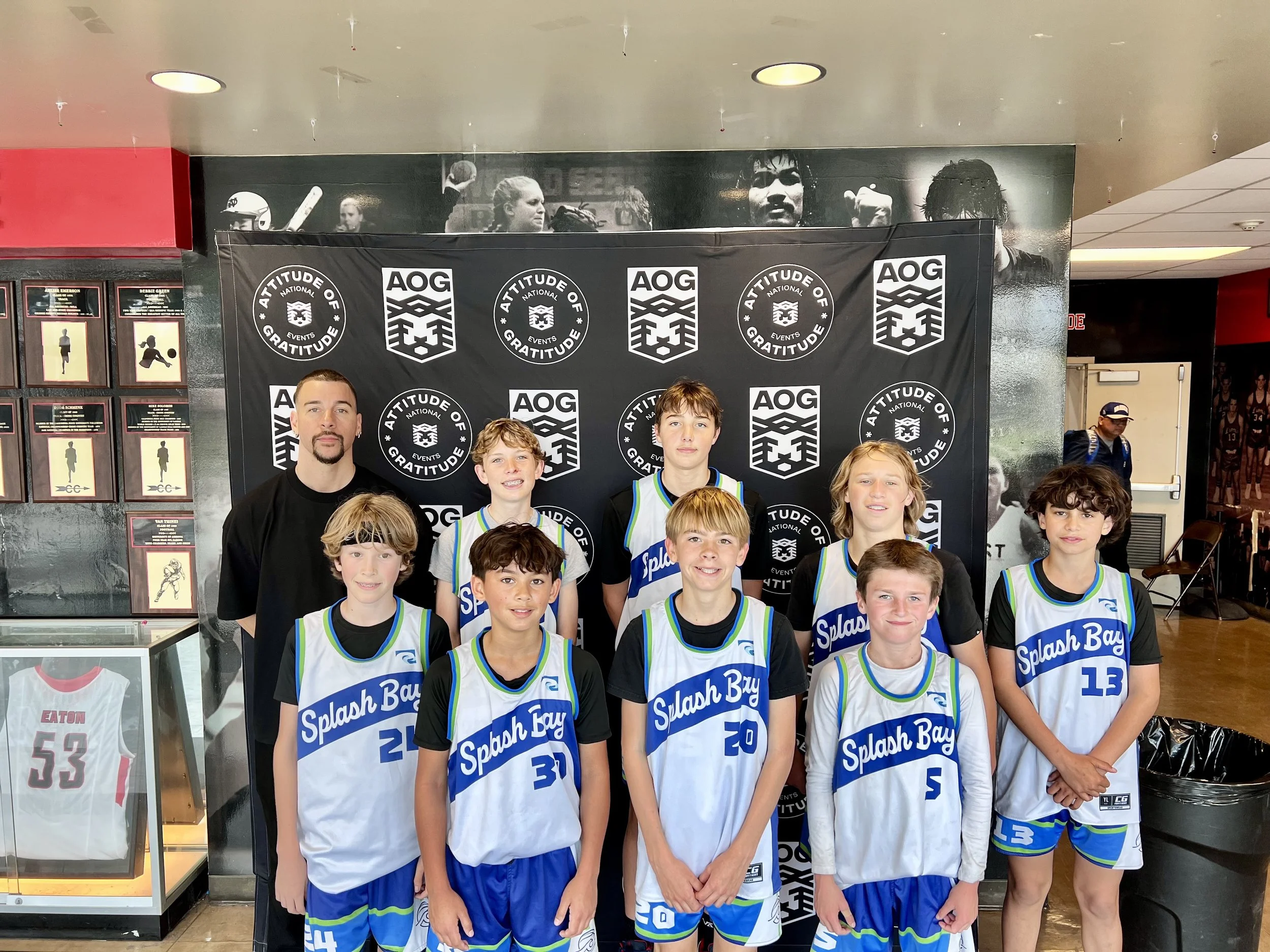 A youth basketball team in white uniforms with blue and green accents, standing with their coach in front of a black backdrop with logos for 'Attitude of Gratitude' and 'AOG' at a sports event.