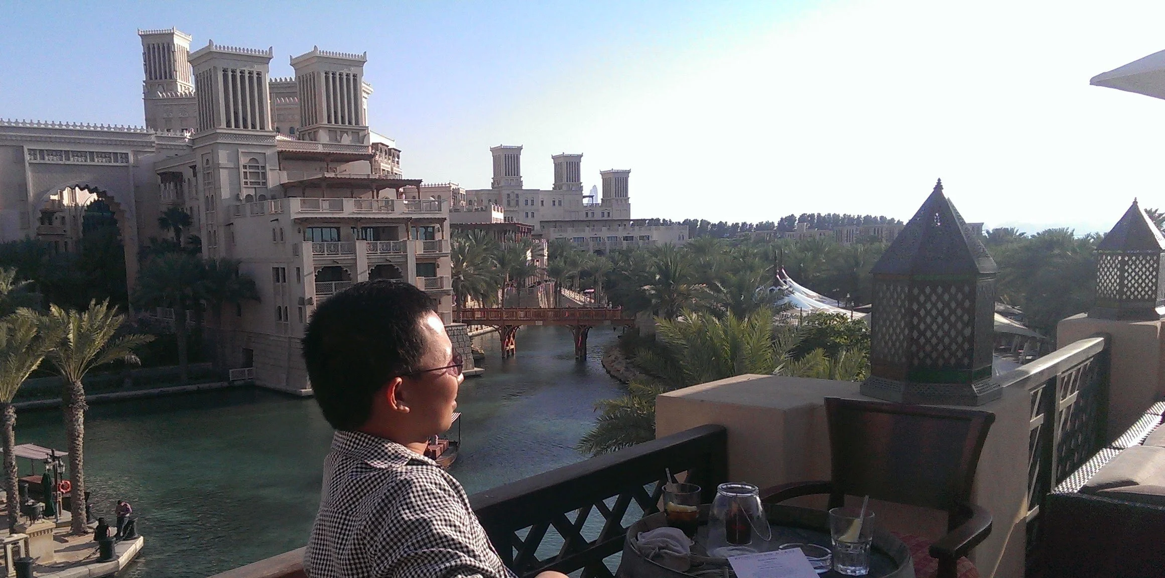 A woman sitting at an outdoor patio overlooking a waterway with palm trees, bridge, and a large white building with towers in the background.