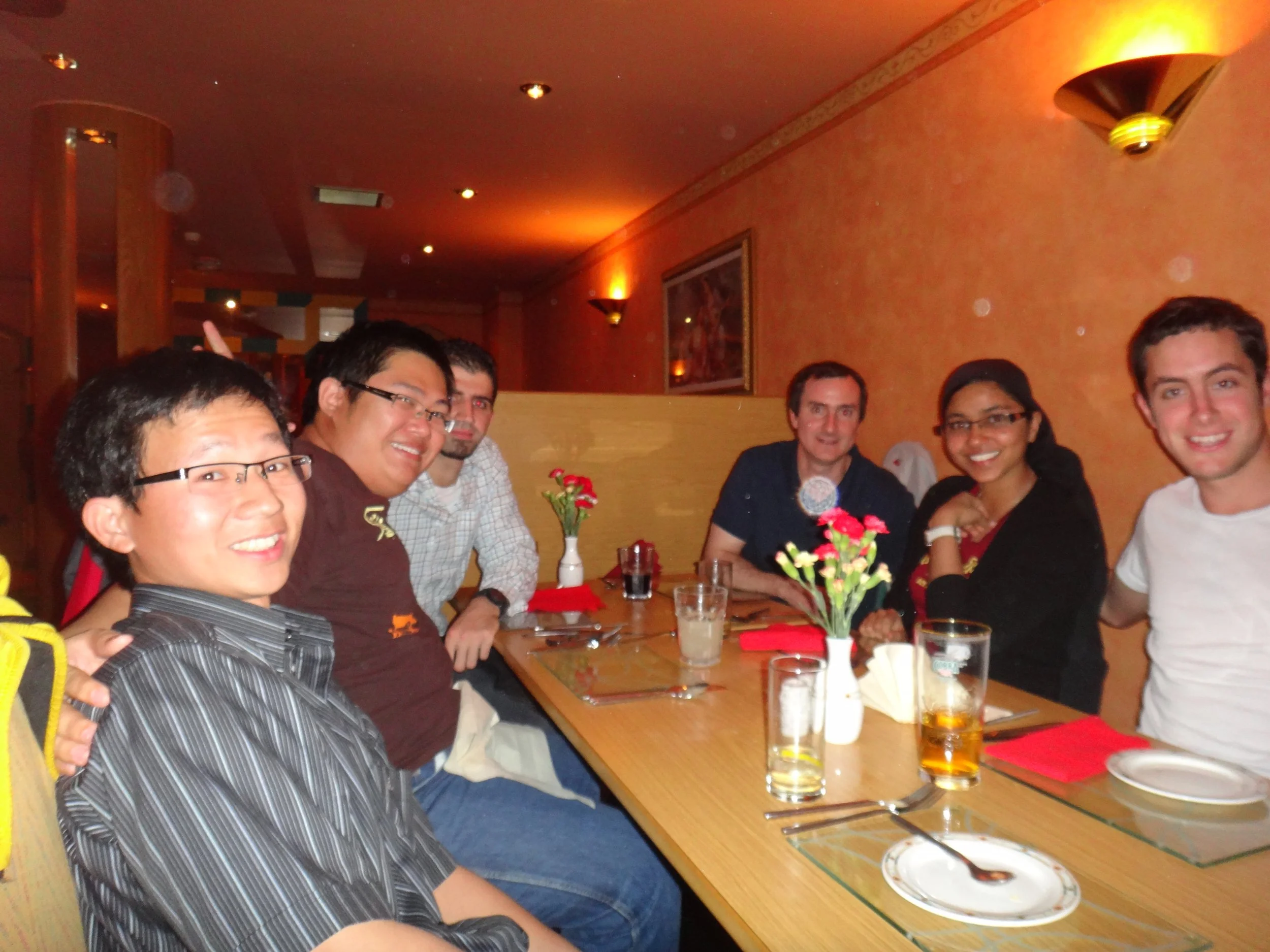 Six people sitting around a dining table in a restaurant, smiling at the camera, with flowers and drinks on the table.