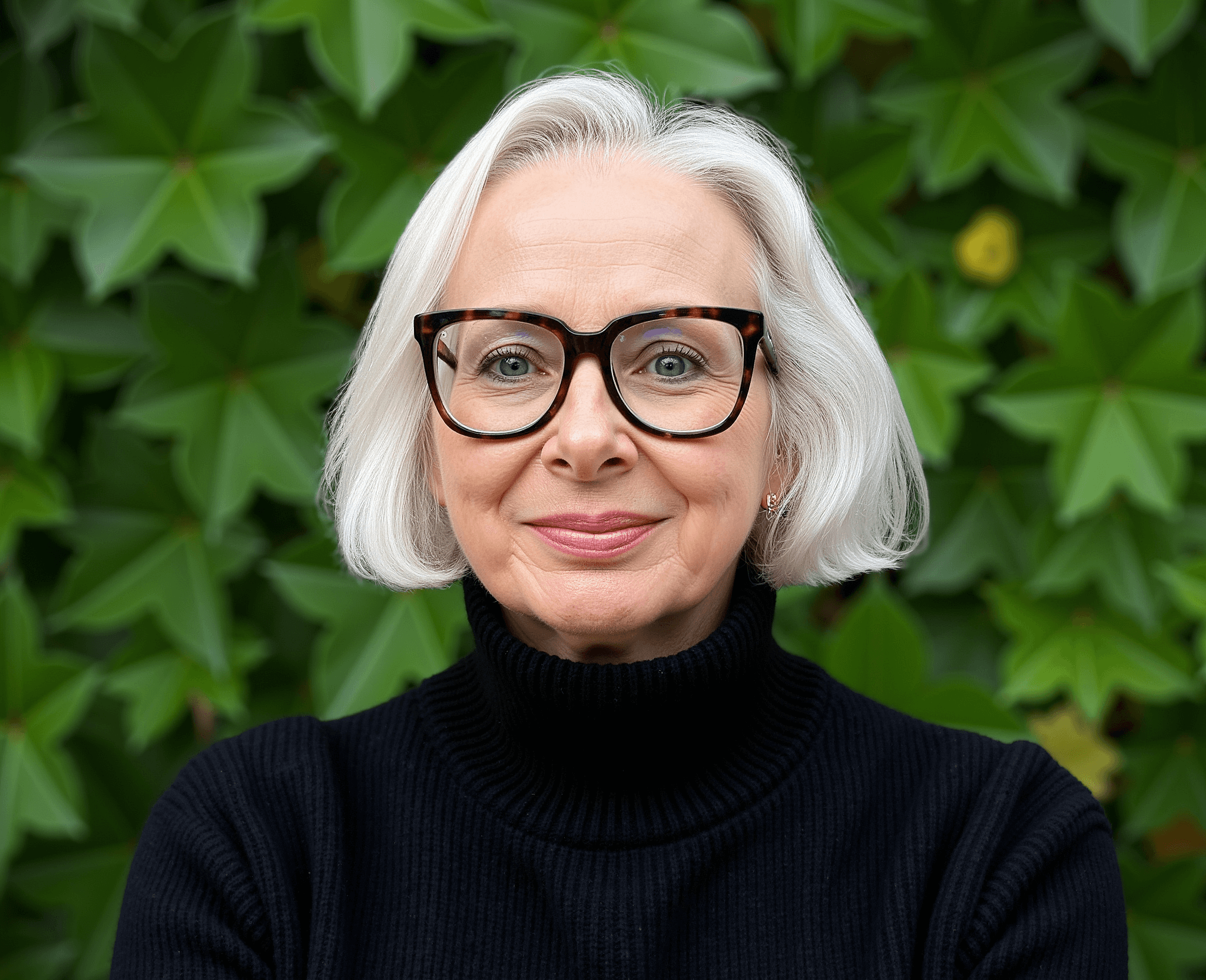 woman wearing black jumper and black glasses standing in front of a hedge in a garden