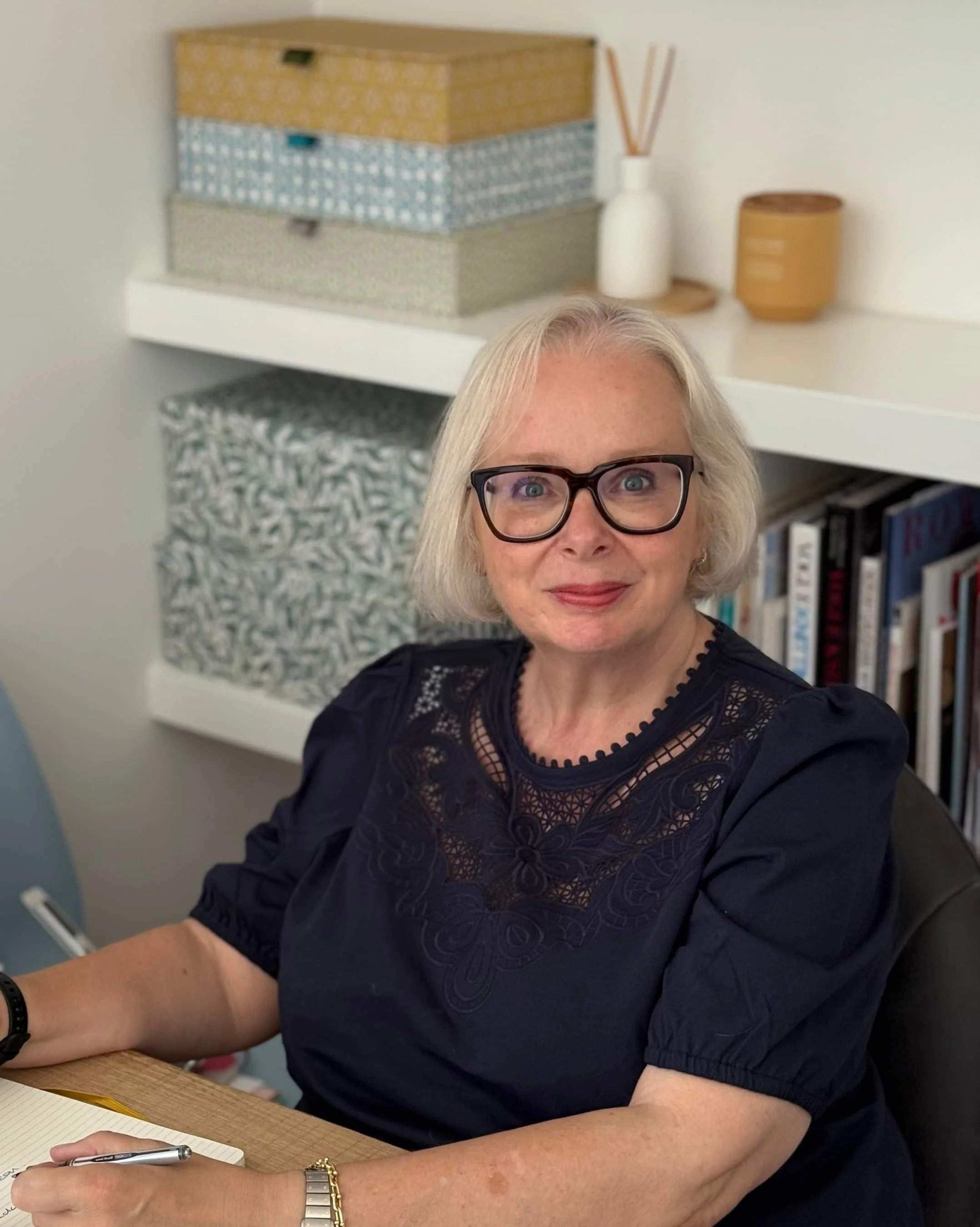 woman wearing black blouse and black glasses sitting in an office with shelves in the background