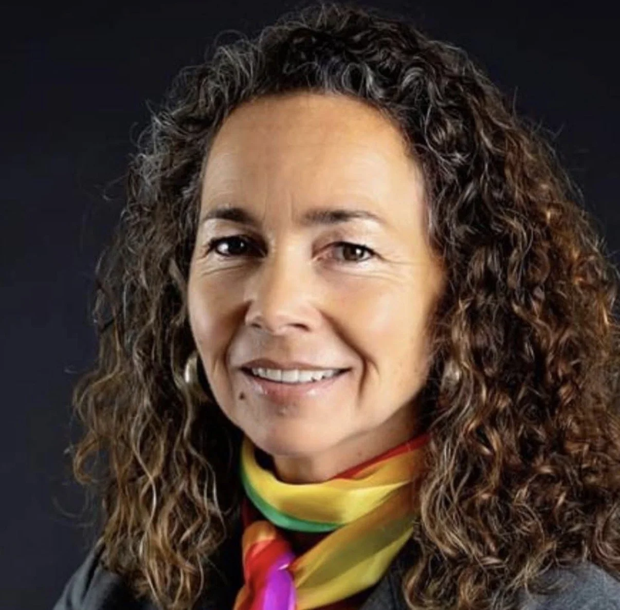 A woman with light skin and curly brown hair smiling outdoors with a blurred green background.