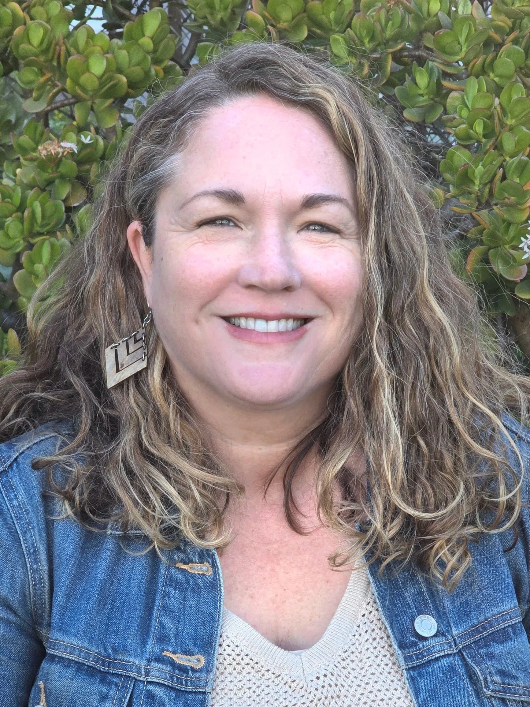 A smiling woman with curly blonde hair, wearing a black jacket and a light-colored scarf, outdoors with greenery in the background.