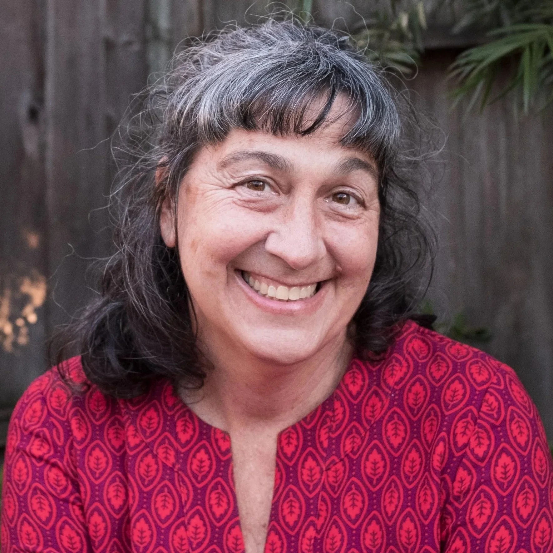 A woman with light skin and salt-and-pepper hair smiling outdoors, wearing a red patterned top, in front of a wooden fence and green foliage.
