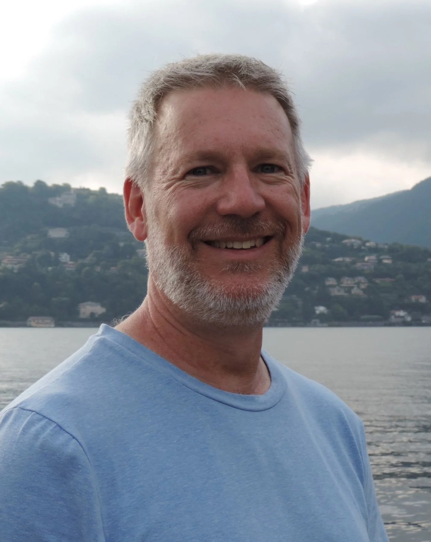 Smiling man with short gray hair and beard, wearing a light blue shirt, standing near a body of water with hills and houses in the background under cloudy sky.