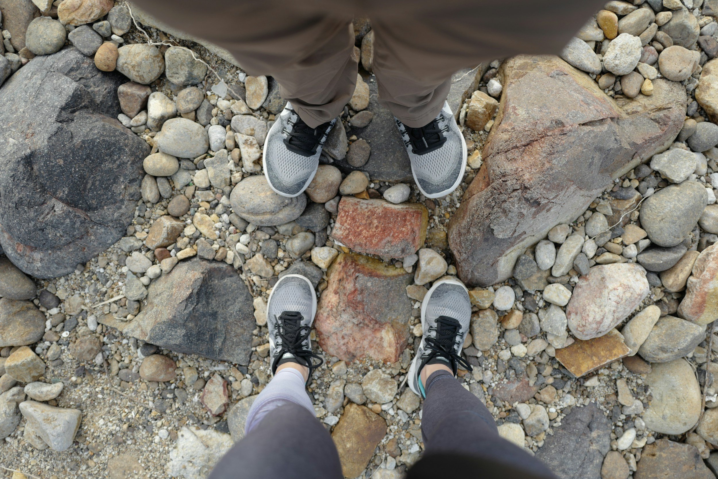 A top-down view of two people standing on a rocky beach, showing their feet and legs. One person is wearing gray athletic shoes and black leggings, and the other is wearing gray athletic shoes and khaki shorts.