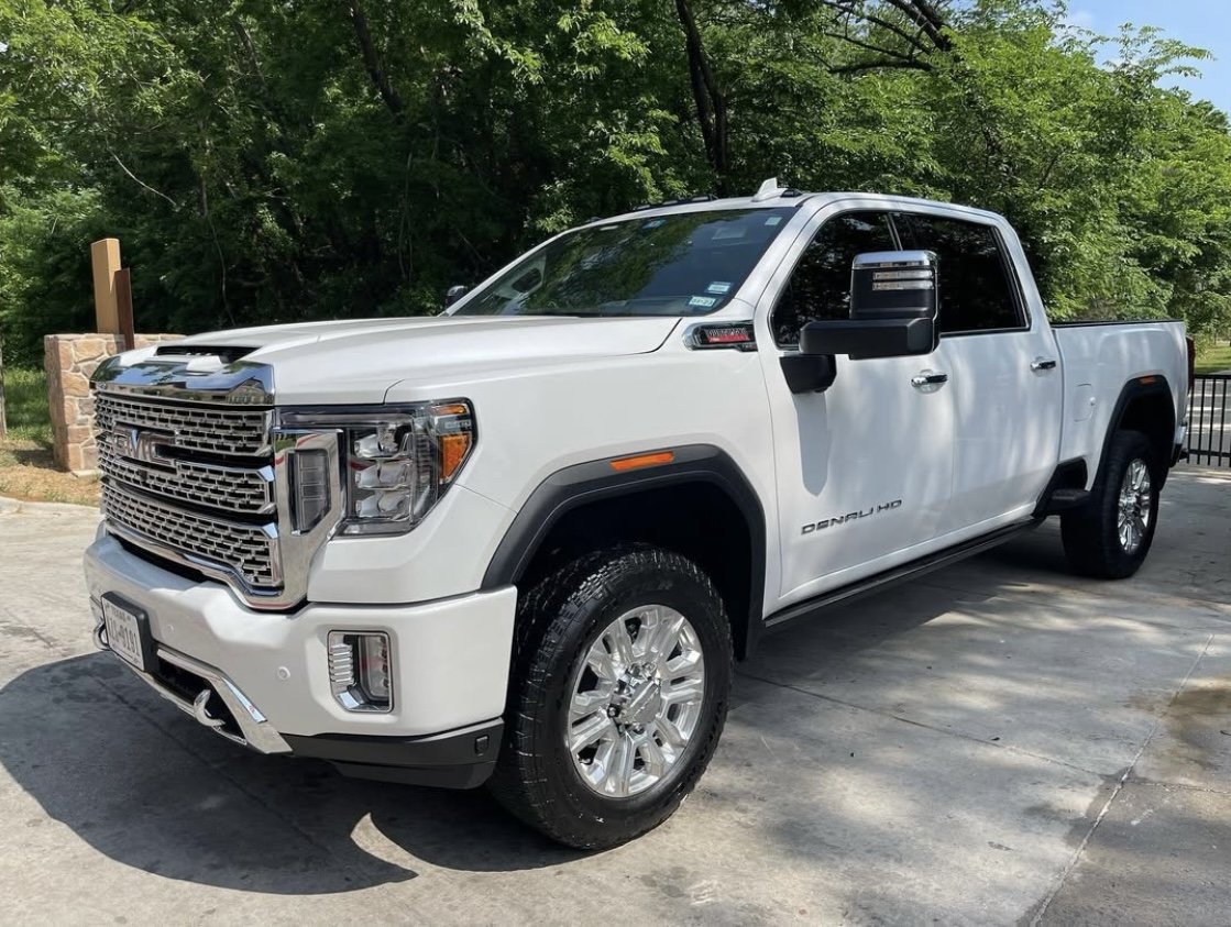 A white GMC Sierra Denali HD pickup truck parked on a concrete driveway, with trees and a stone column in the background.