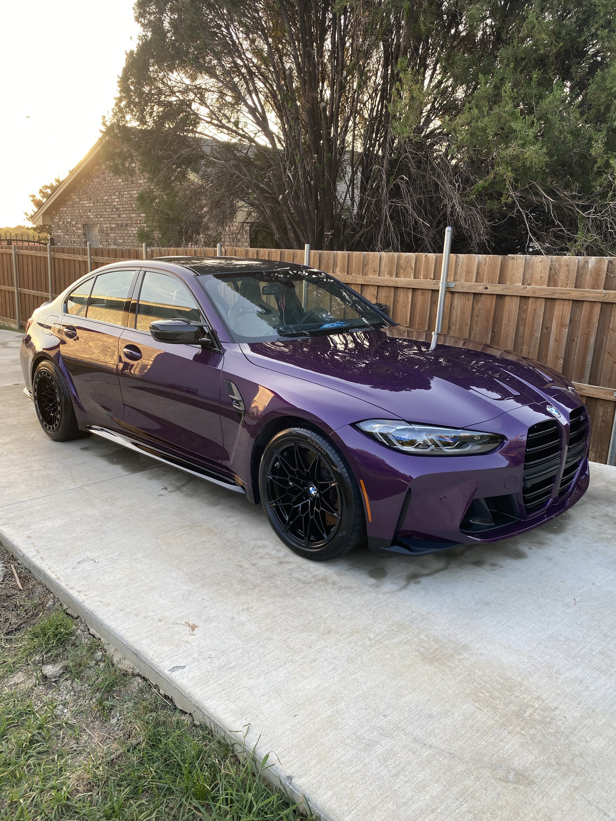 A purple BMW sedan with black wheels parked on a concrete driveway, with a wooden fence and trees in the background, during sunset.