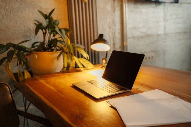 Open laptop, stack of papers, desk lamp, and potted plant on a wooden desk in a cozy room.