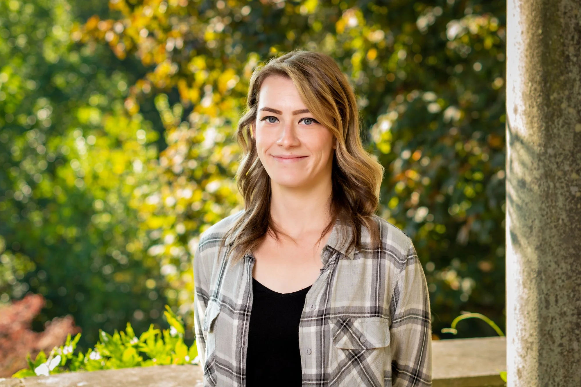 A young woman with wavy brown hair smiling outdoors in front of a background of trees and foliage, wearing a plaid shirt over a black top.