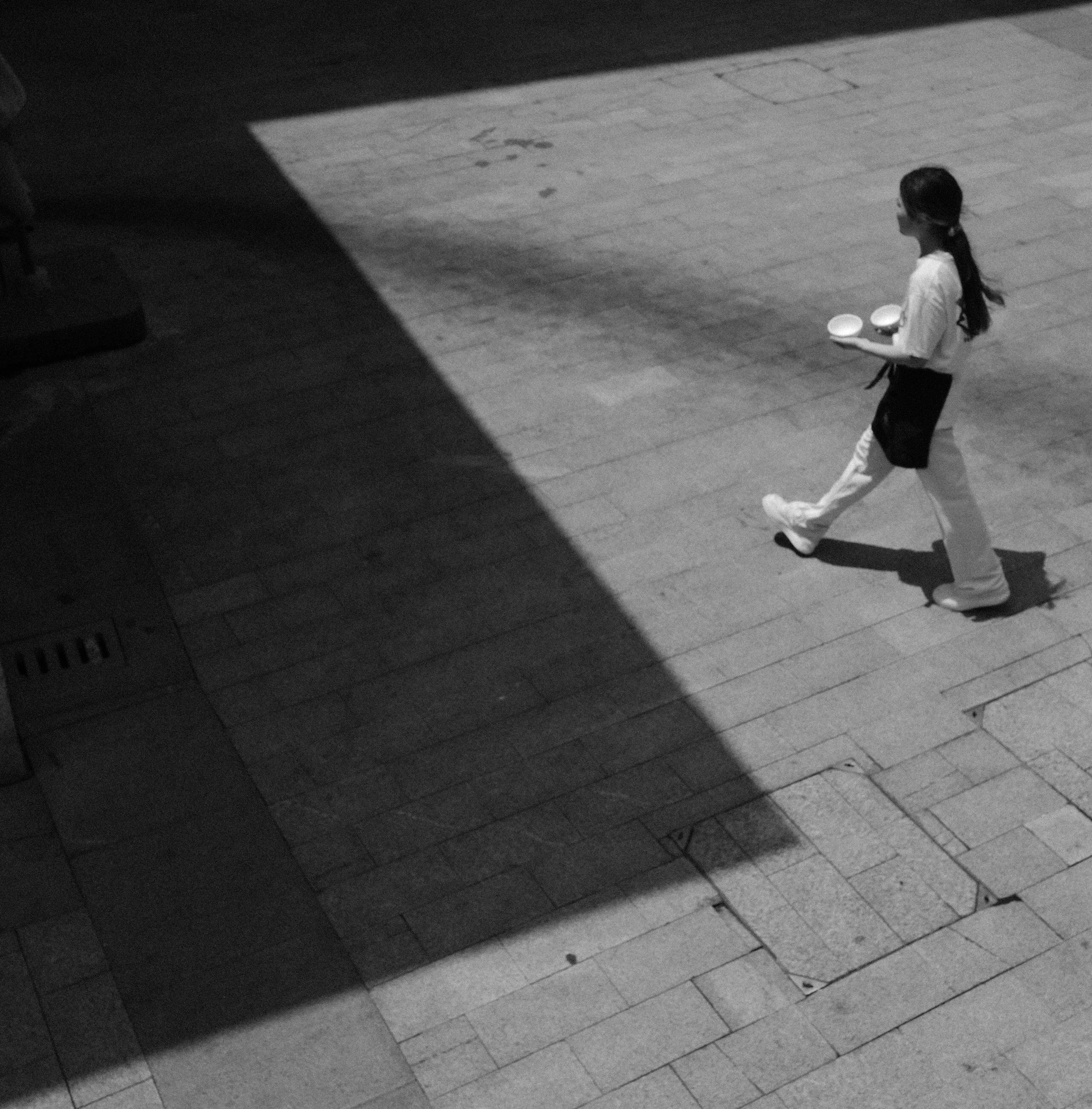 The Path: A waitress crosses a sunlit square, with a strong play of midday shadows