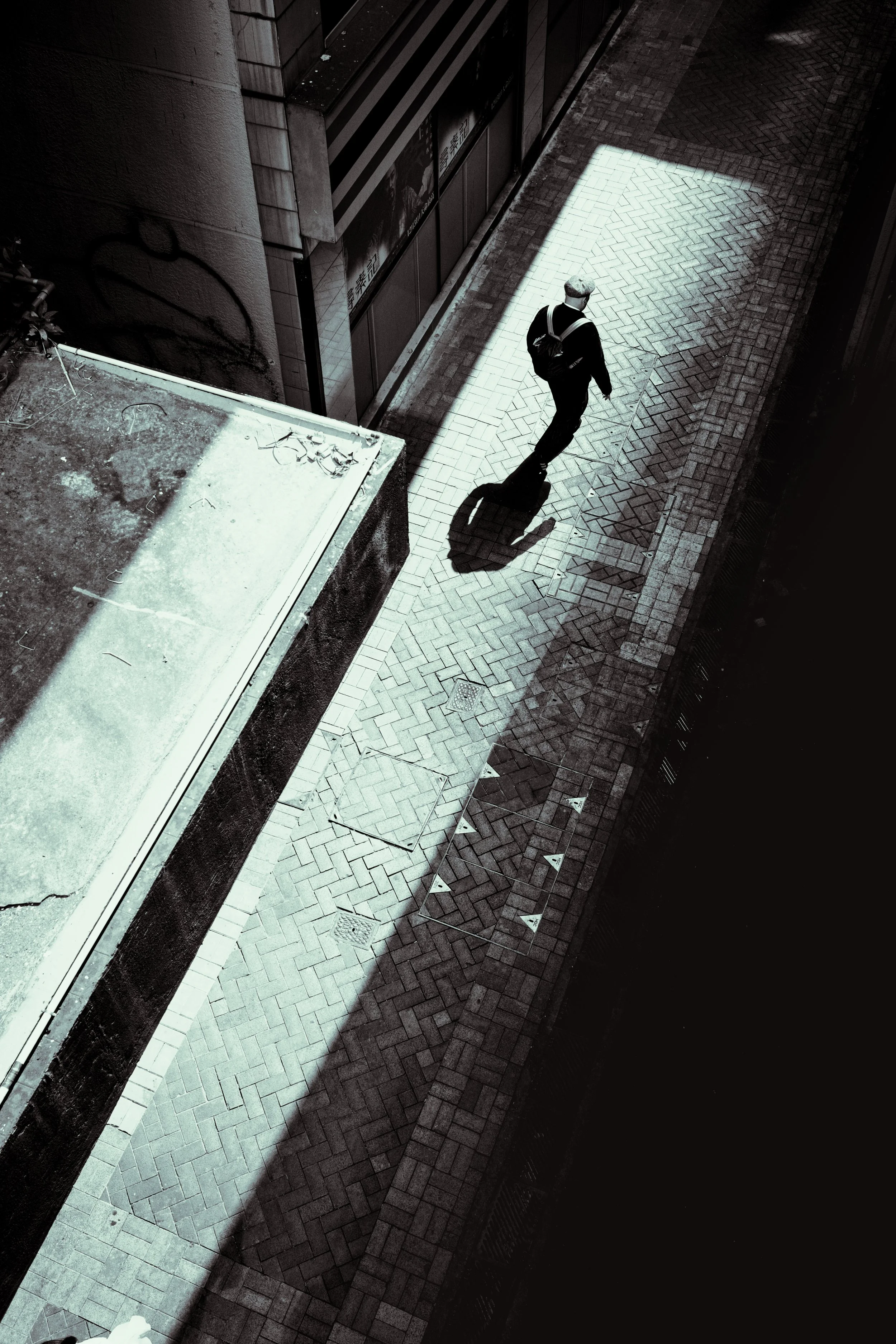 A person with gray hair walking on a narrow brick sidewalk, illuminated by sunlight, with shadows cast by nearby buildings, and a wall with graffiti on the left.