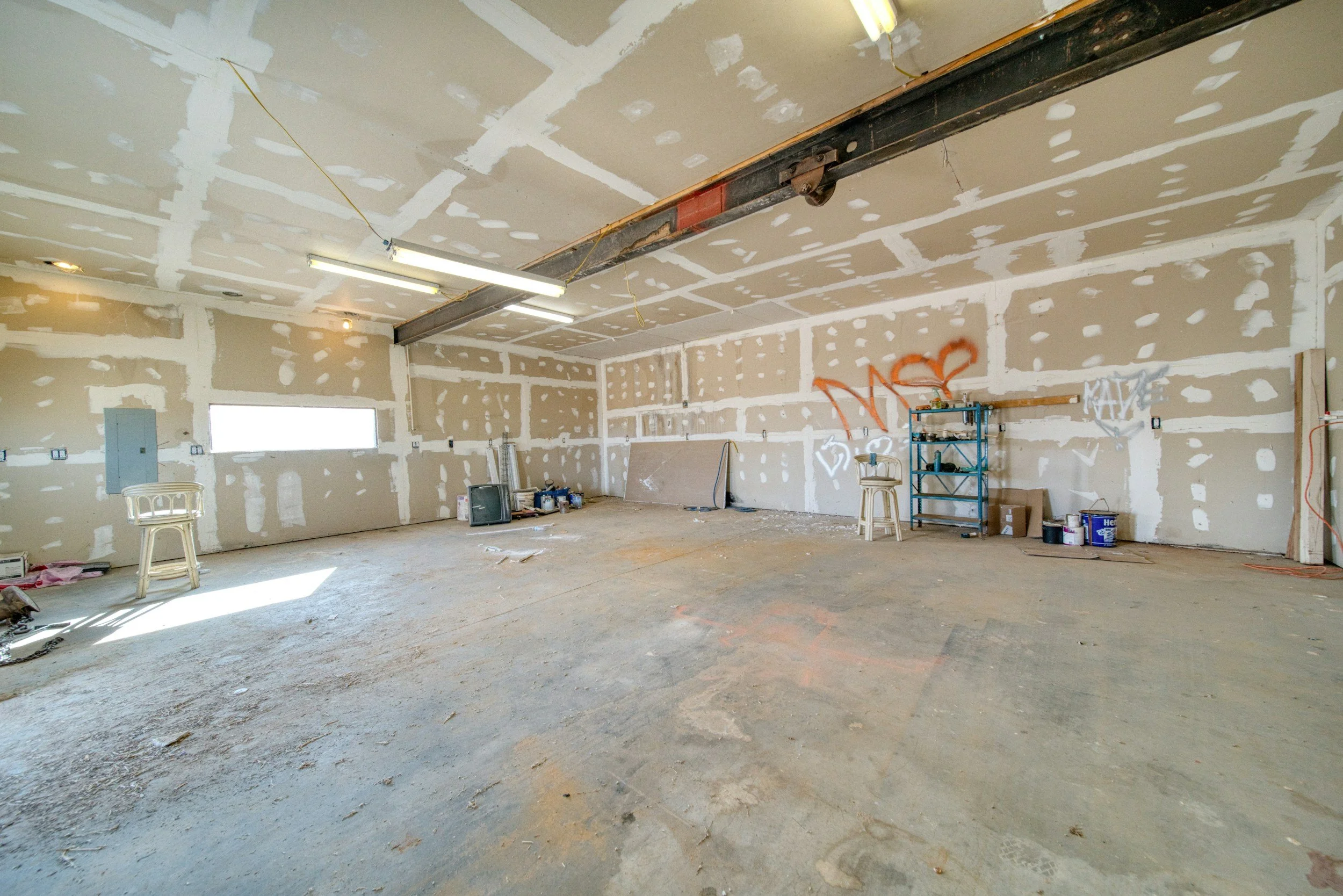 Empty garage with unfinished walls, garage door opener rail on ceiling, a window on the left wall, and some scattered tools and chairs.