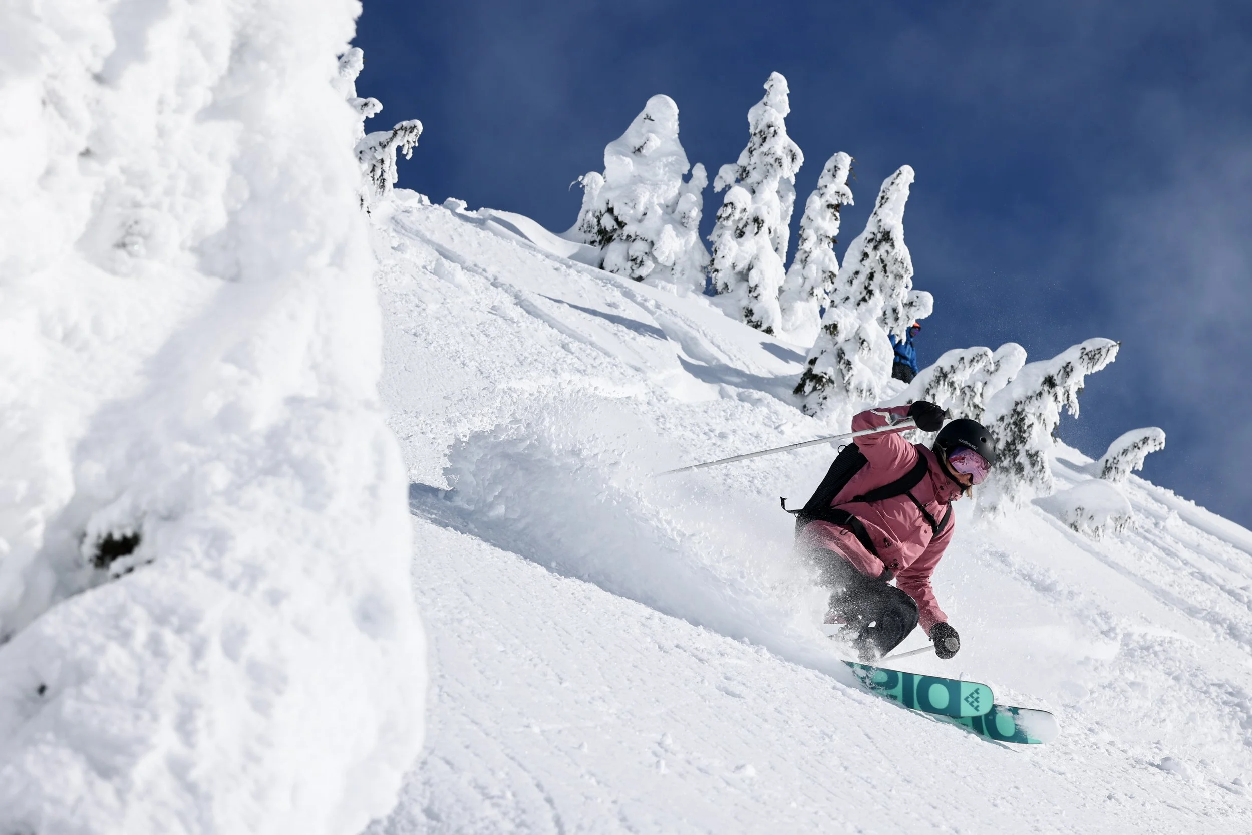 A person in pink ski gear and a helmet skis down a snow-covered mountain slope, surrounded by snow-covered trees, under a clear blue sky.