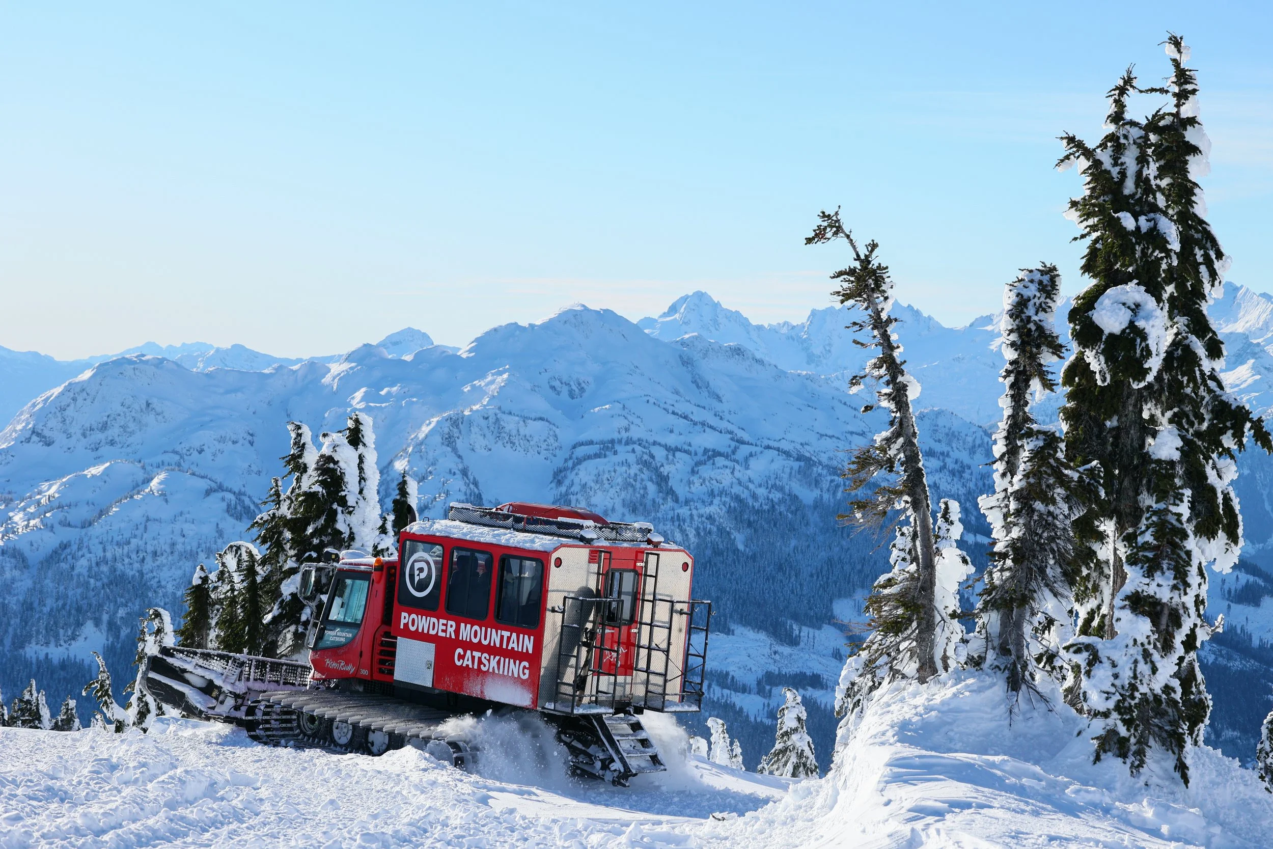 A snowcat vehicle labeled 'Powder Mountain Catskiing' on a snowy mountain slope with snow-covered trees and mountains in the background.