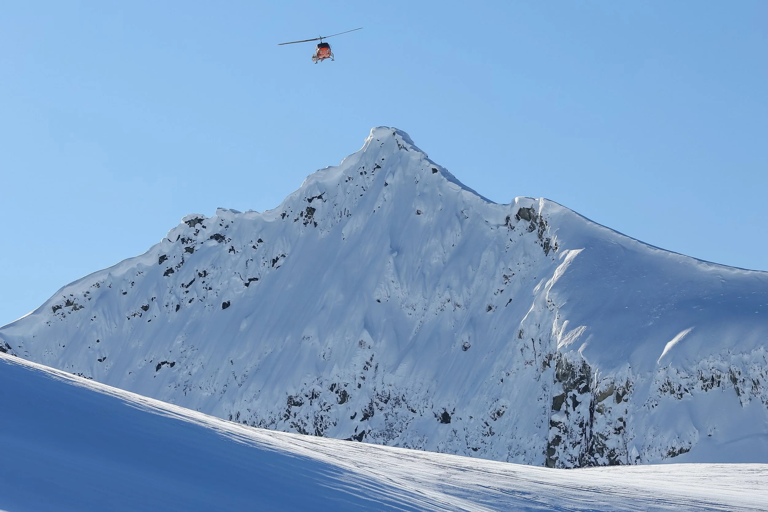 Snow-covered mountain peak with a helicopter flying above against a clear blue sky.
