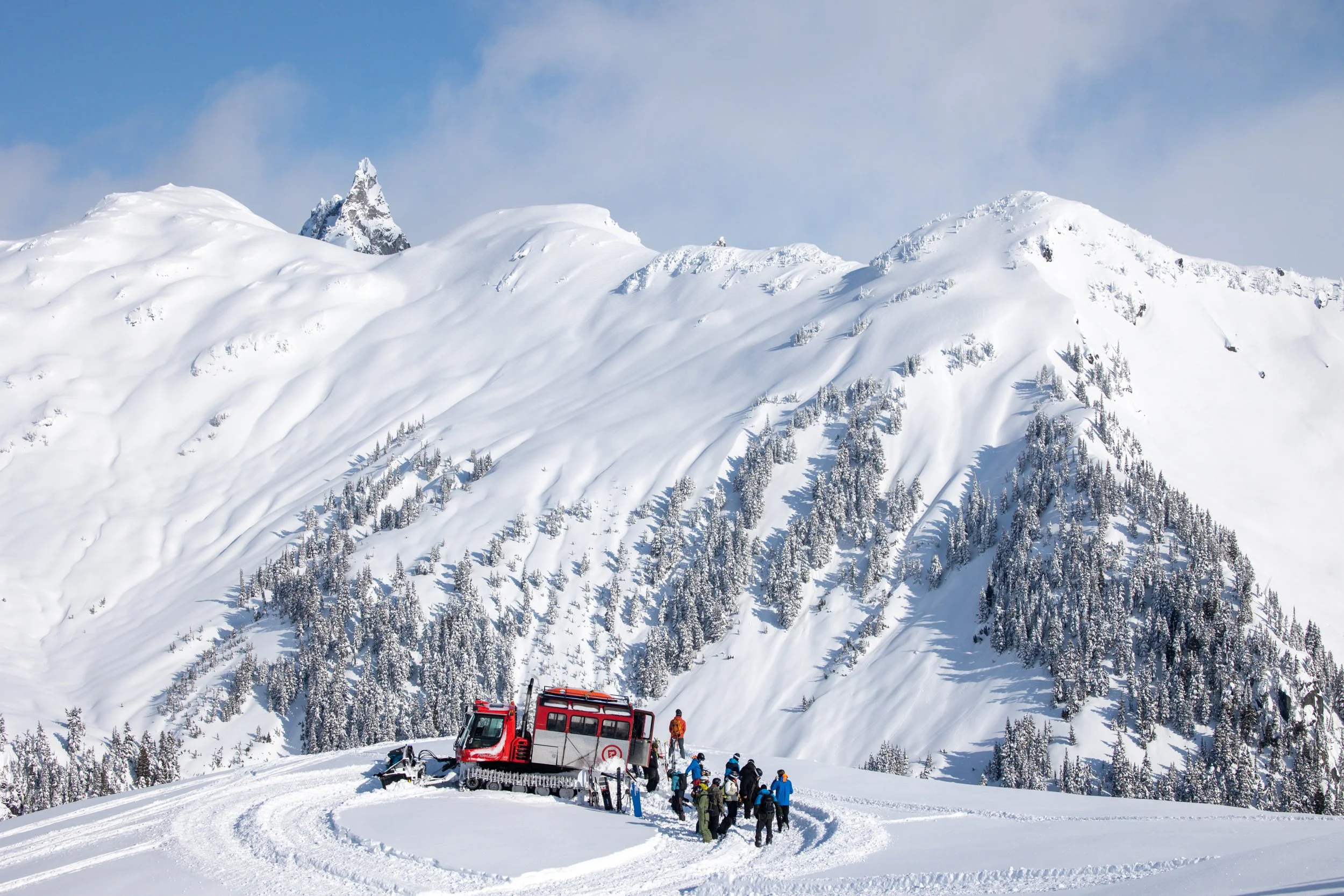 A group of people in winter clothing standing near a red snow grooming machine in a snow-covered mountain landscape with pine trees and a clear blue sky.