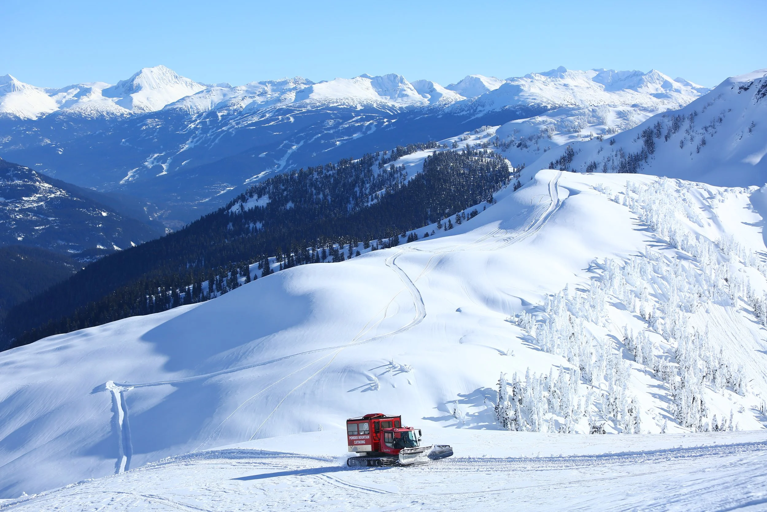 A red snow grooming machine operating on a snowy mountain landscape with snow-covered peaks and trees in the background.