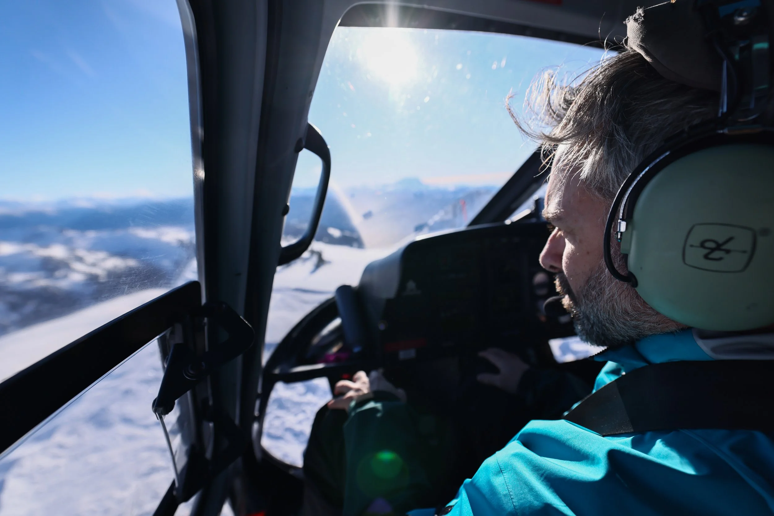 A man with flying in a helicopter over snow-covered mountains, wearing a headset and blue jacket in Whistler