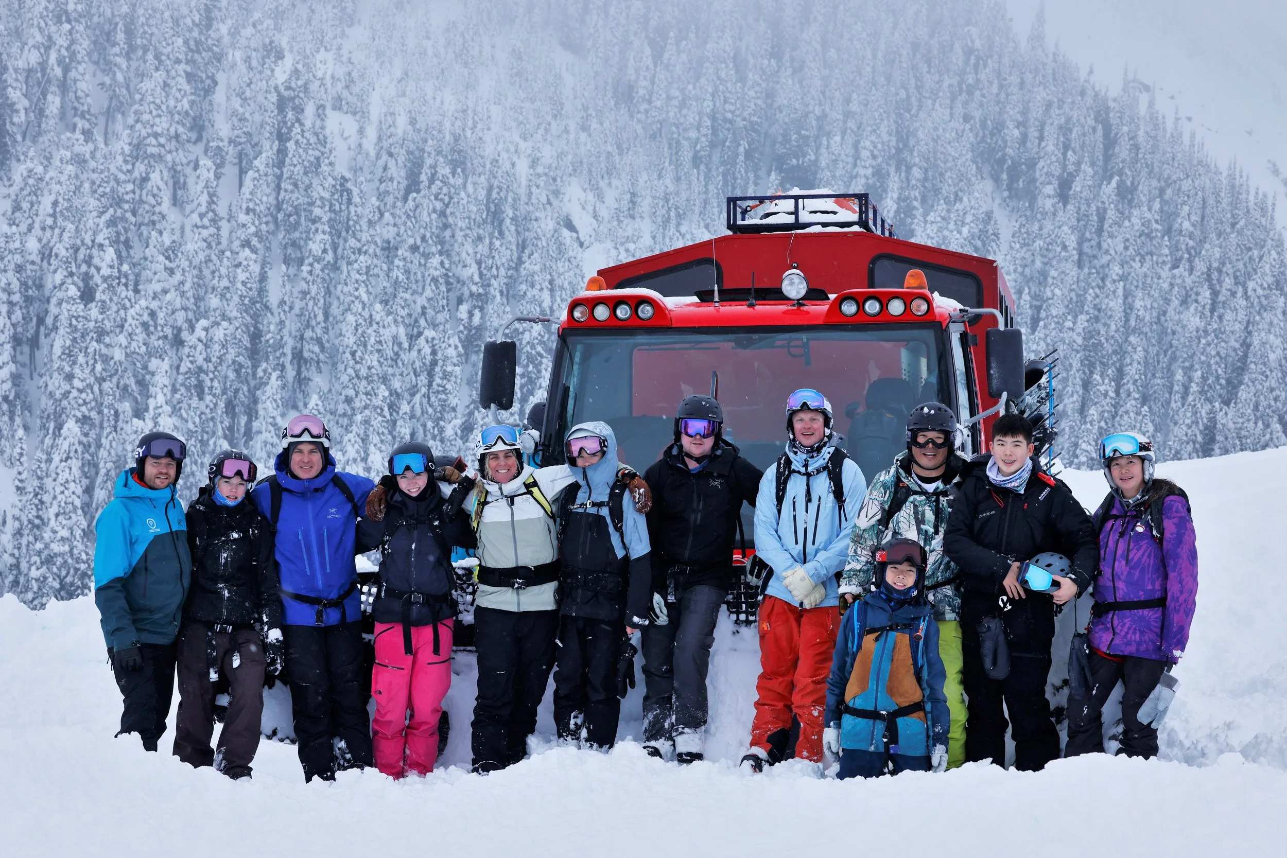 Group of skiers and snowboarders in winter clothing, wearing helmets and goggles, standing in front of a red cat in a snowy mountain landscape.