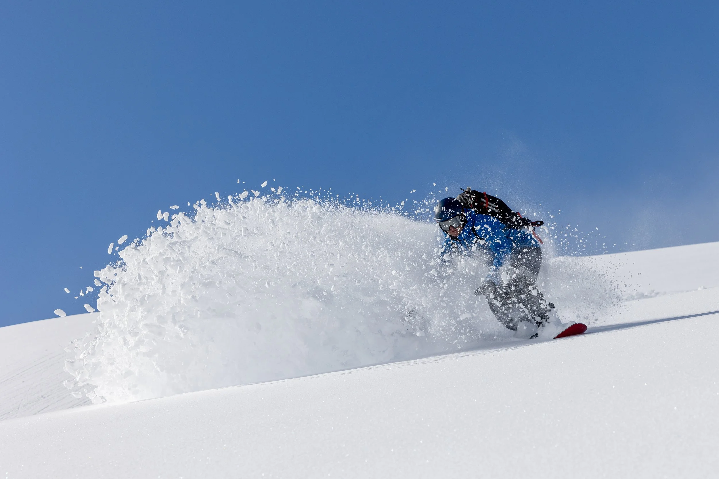 A skier wearing blue jacket and black helmet carving through fresh snow on a mountain slope, creating a spray of snow behind them under a clear blue sky.