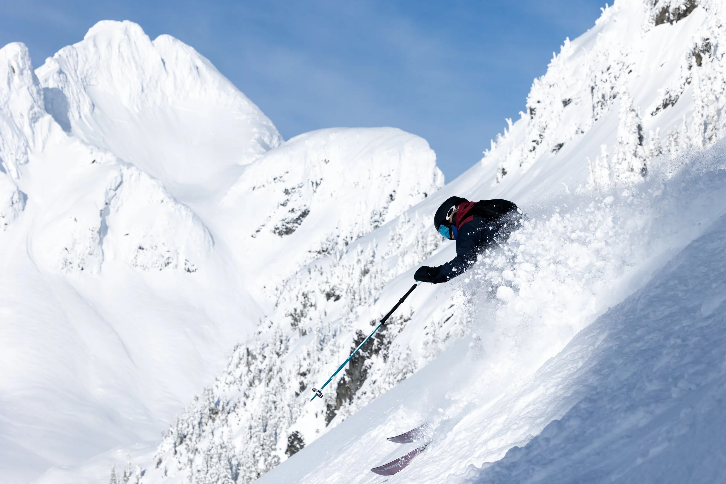 A person wearing a black helmet and black winter gear skiing down a snowy mountain slope with snow-covered peaks in the background.