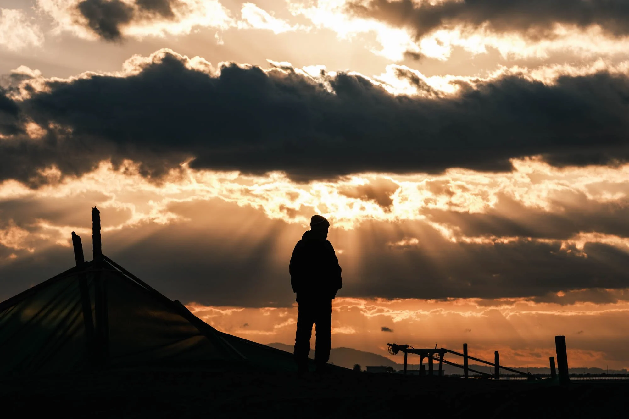 Sunrise bursting through clouds as a lone figure stands in silhouette on a beach in Fujisawa, Japan.