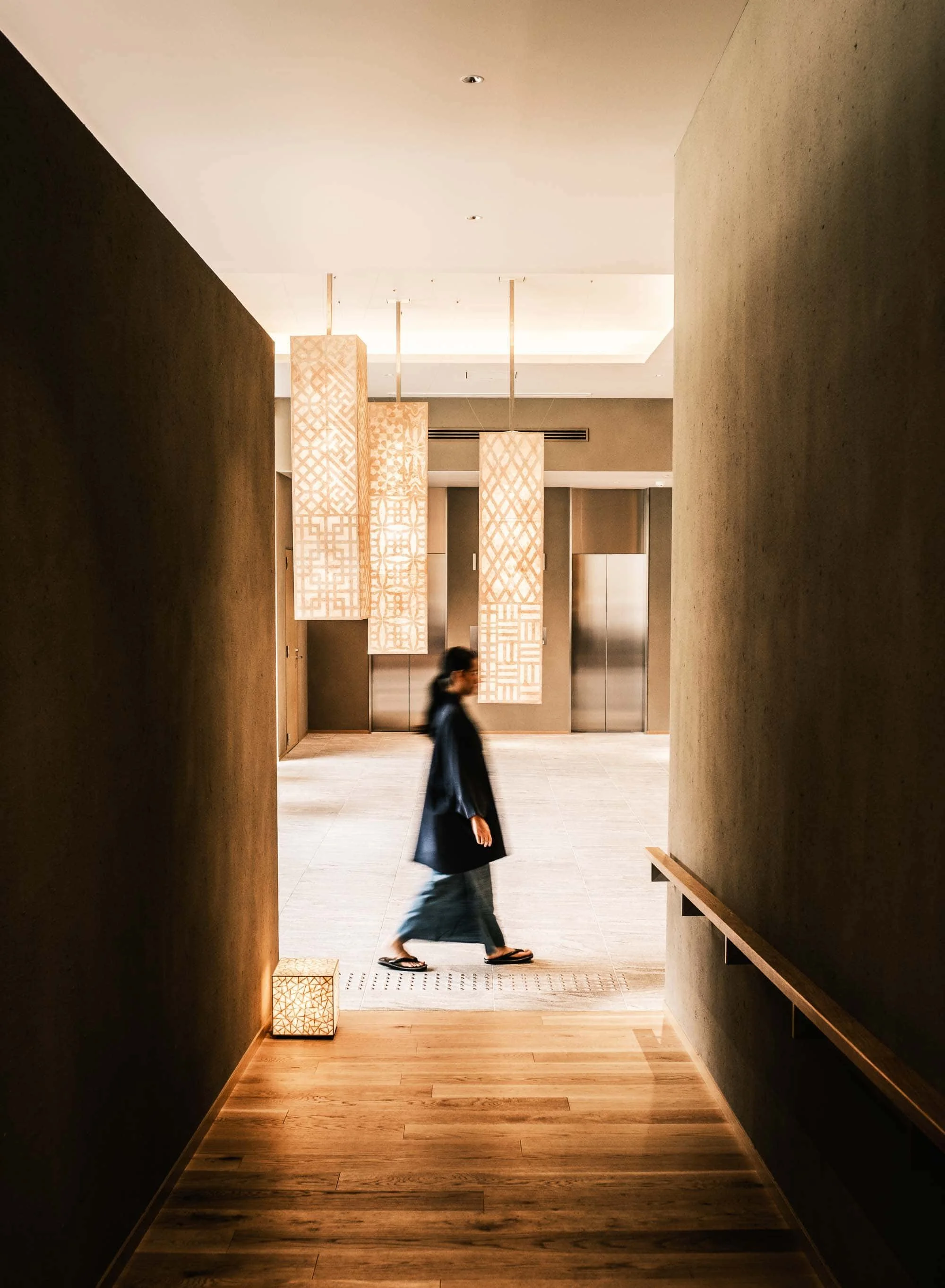A woman wearing traditional Japanese clothing called yukata walking in a modern hotel lobby with wooden accents, hanging decorative lanterns, and elevators in the background.