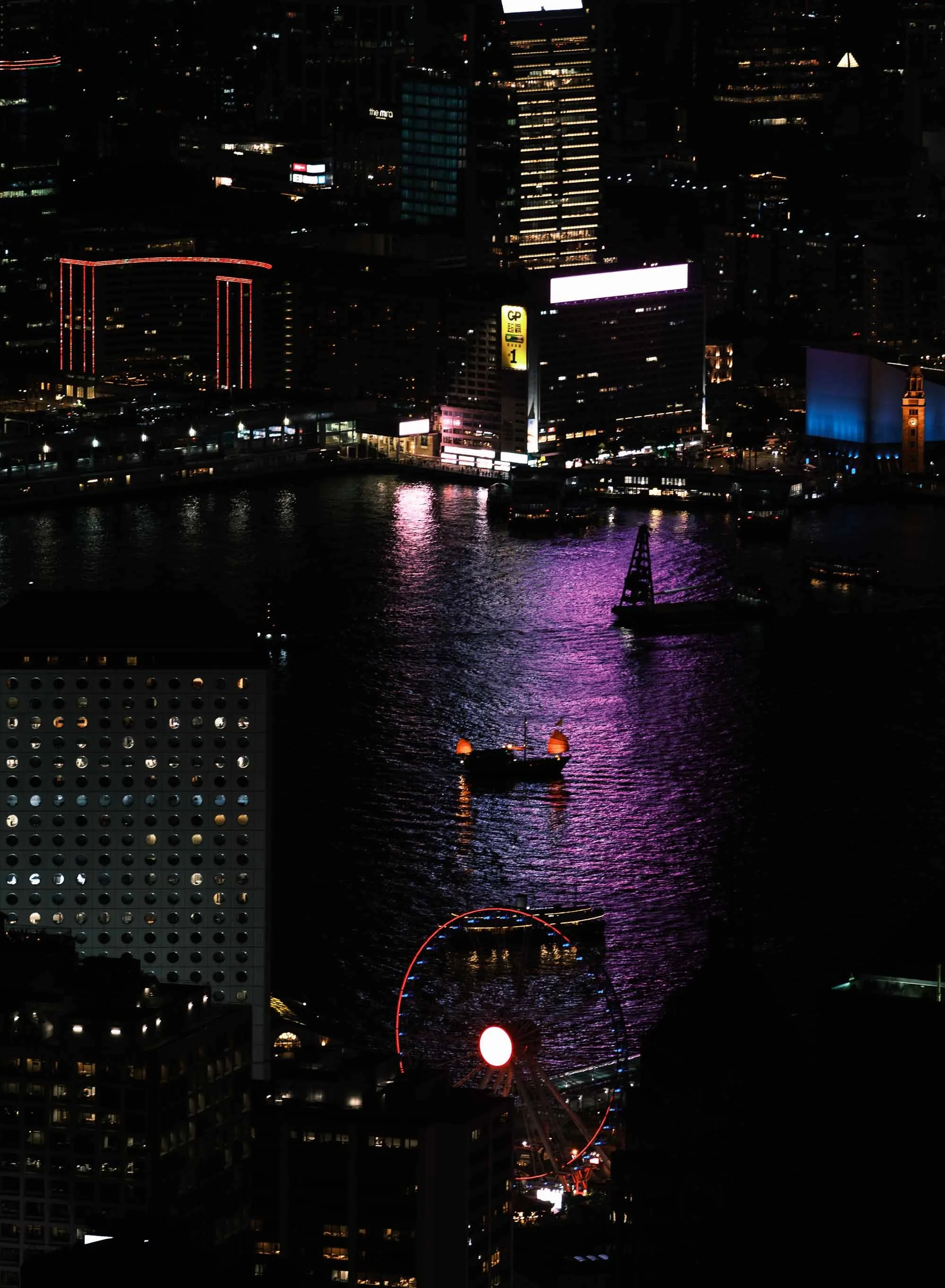 Nighttime cityscape of Victoria Harbour Hong Kong, with the lit-up red sails of the Aqua Luna cruise boat on the water.