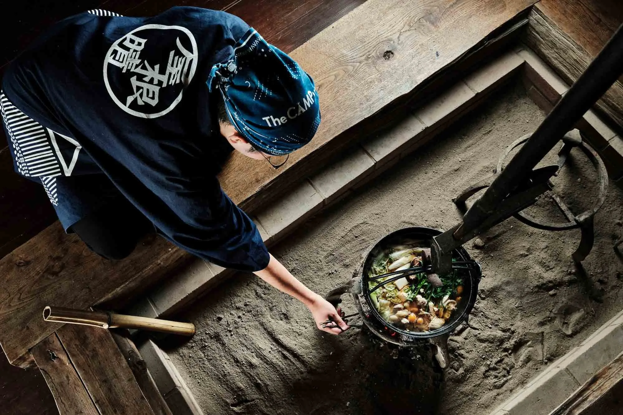 A man in traditional Japanese attire cooking soup over an open floor hearth with a pot suspended by a chain from the ceiling.