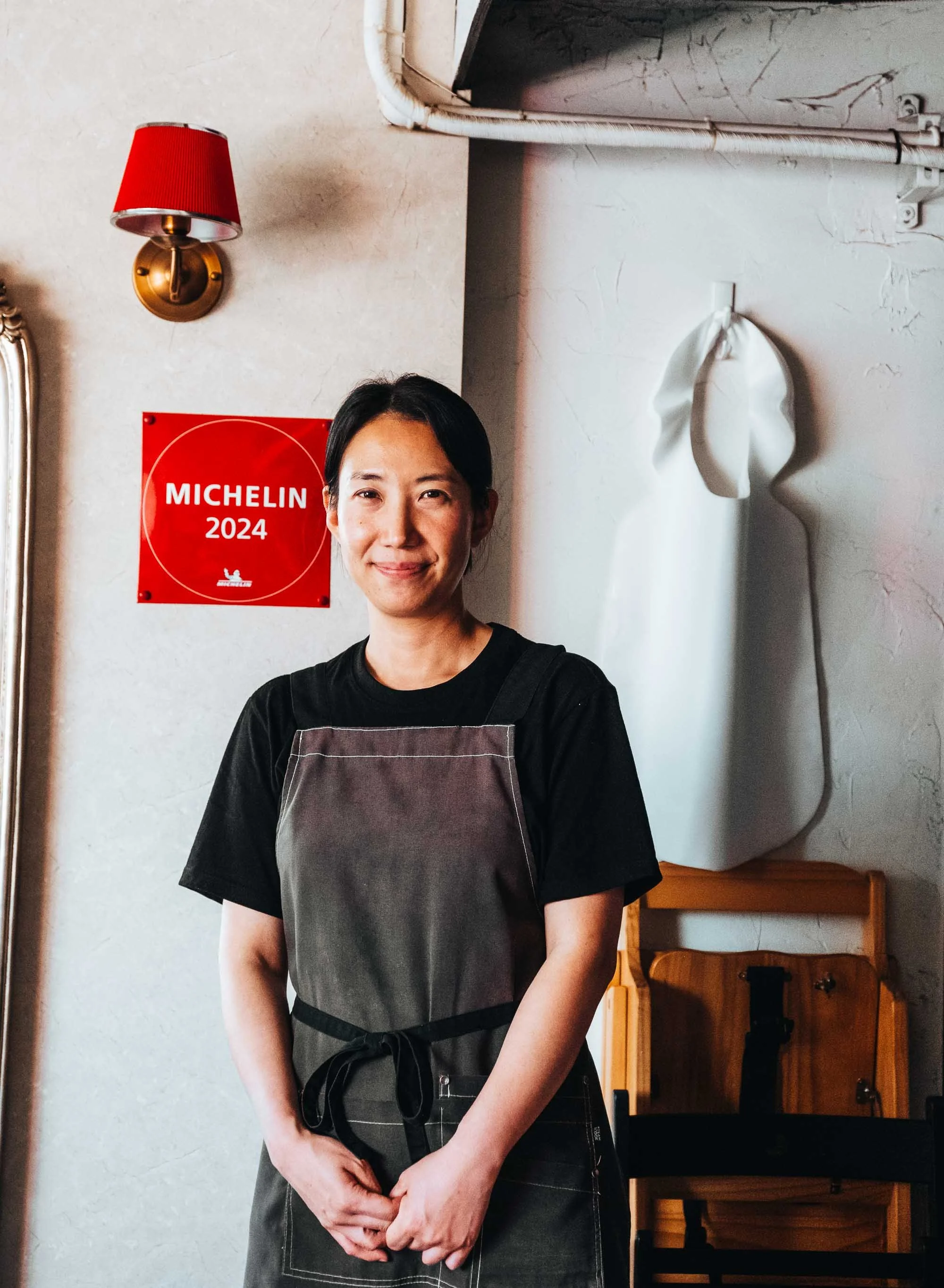 A Korean restaurant owner standing inside her Busan ramen shop next to a red Michelin sign.