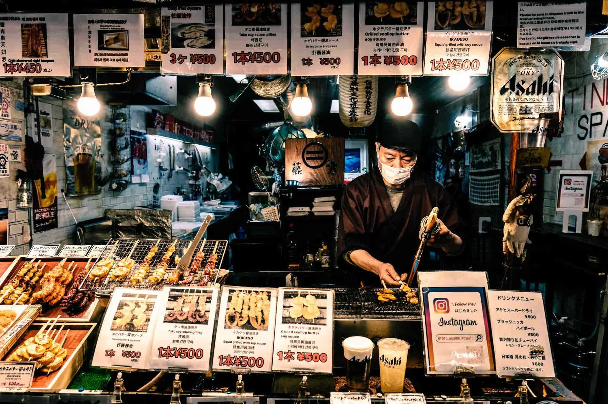 A man wearing a face mask preparing skewers of grilled seafood at a Kyoto street food stall, with various skewers on display and colorful signage above.