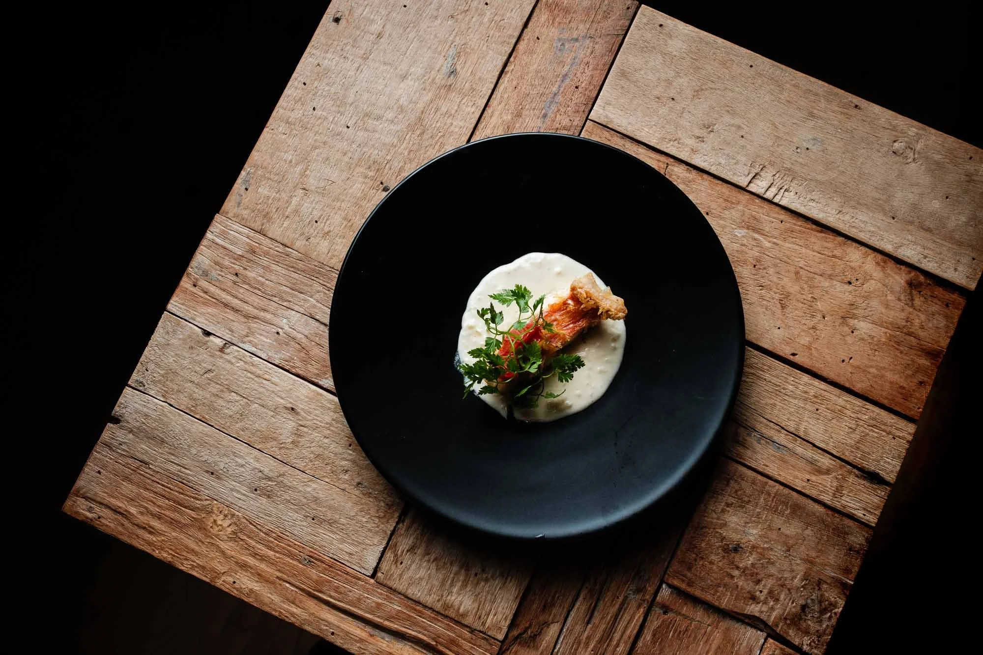 A black plate with a piece of fried chicken on a bed of creamy white sauce garnished with fresh herbs, placed on a rustic wooden table.