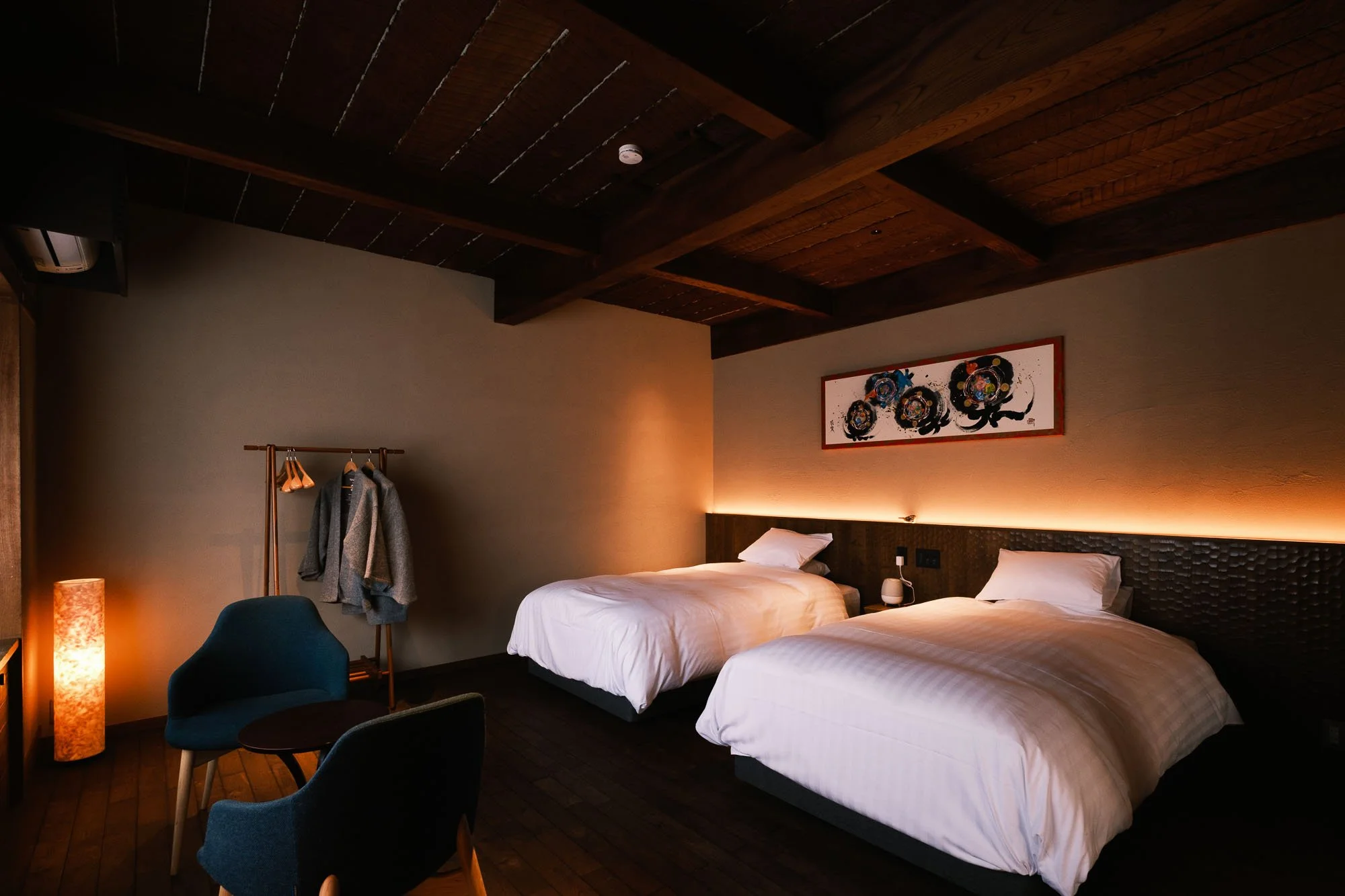 Hotel room with two beds, a dark wood ceiling, modern wall art, a sitting area with two blue chairs and a small table, and a coat rack with jackets.