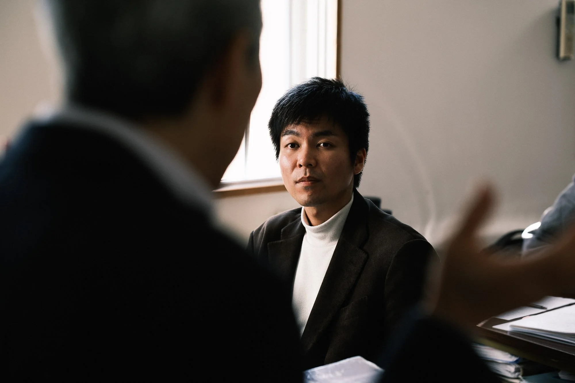 A man with black hair and a white turtleneck shirt sits in a classroom looking a professor. Sunlight streams through a window, illuminating the scene.