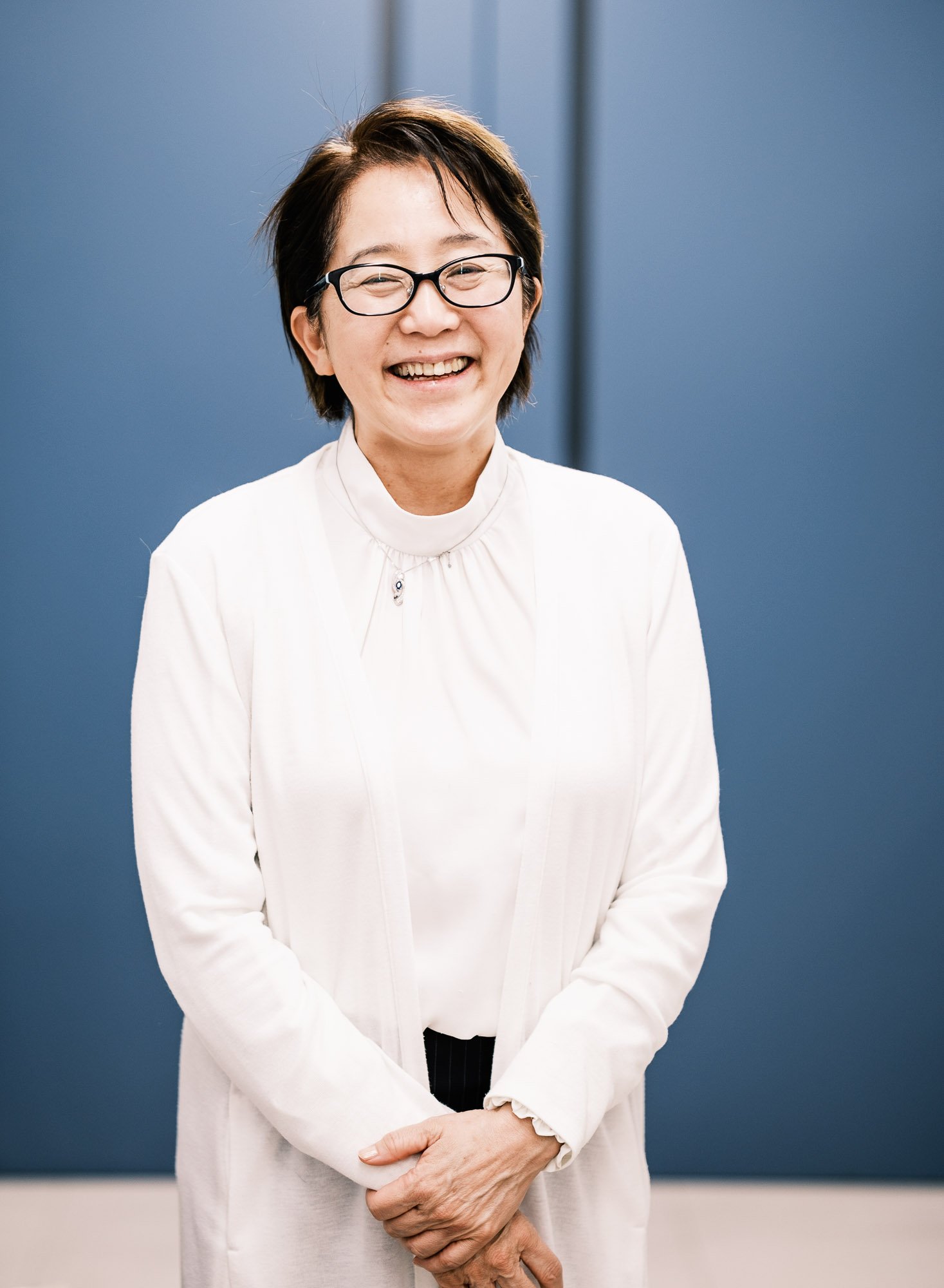 Editorial portrait of a female science researcher with short dark hair, glasses, smiling, wearing a white blouse and jacket, standing in front of a blue wall.