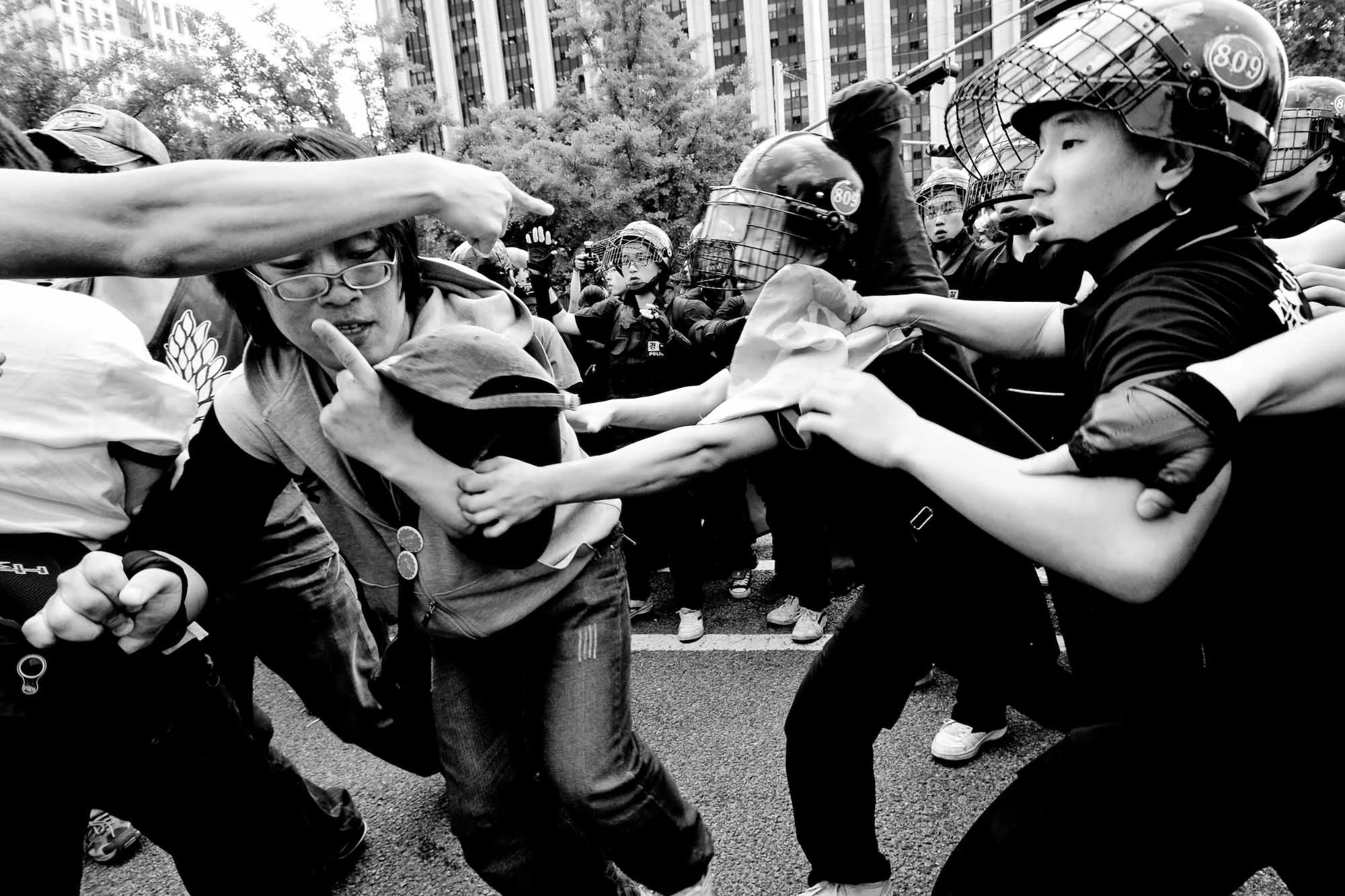 Black and white photo of a street protest with police in riot gear confronting a civilian. The civilian is being pushed or grabbed by police officers, with some officers holding shields and wearing helmets with visors.
