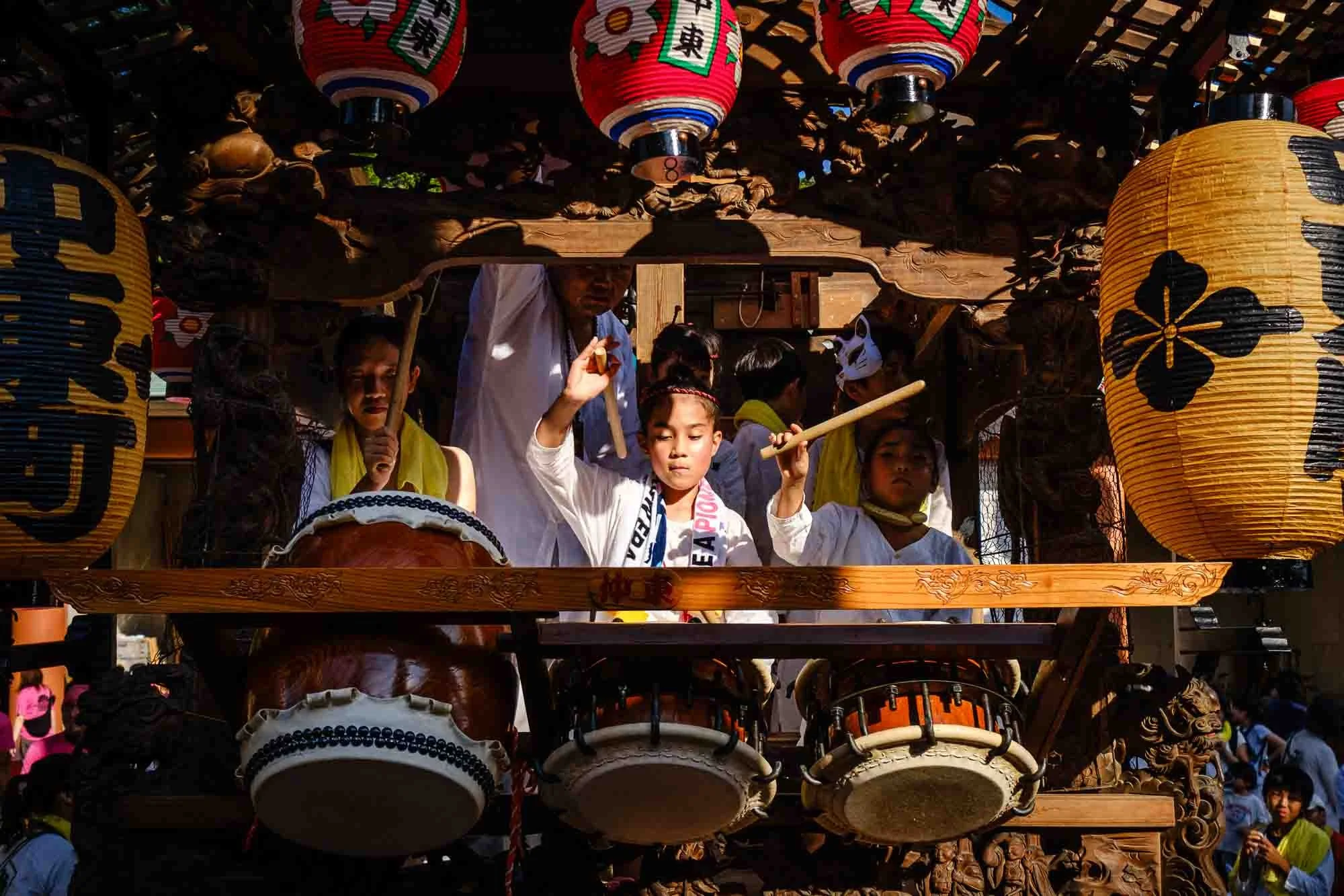 A woman and two girls in traditional clothing play drums on a wooden float at a Japanese summer festival.