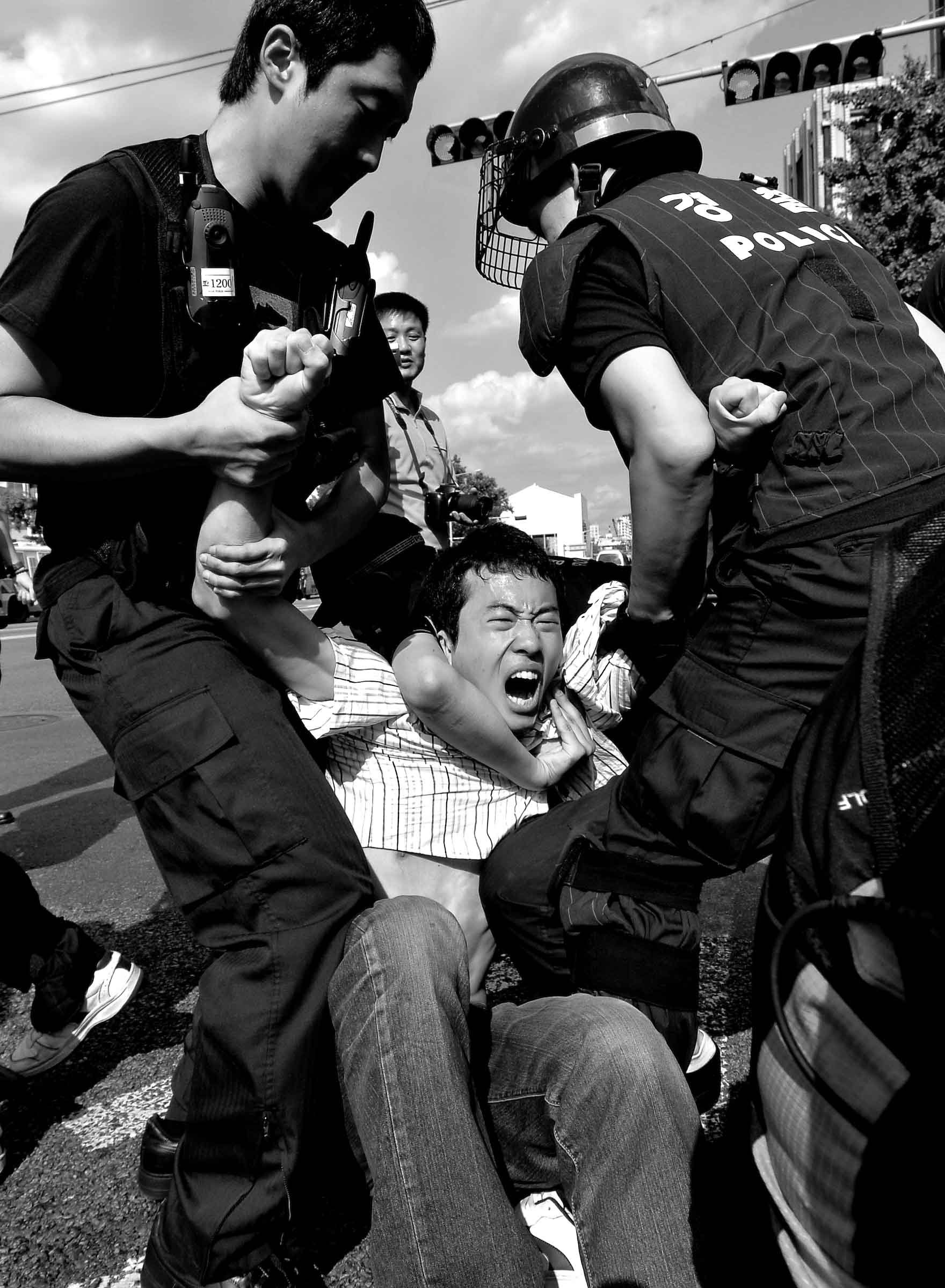 A person is being detained by police officers during a protest. The detained individual appears distressed as police officers hold them down on the ground.