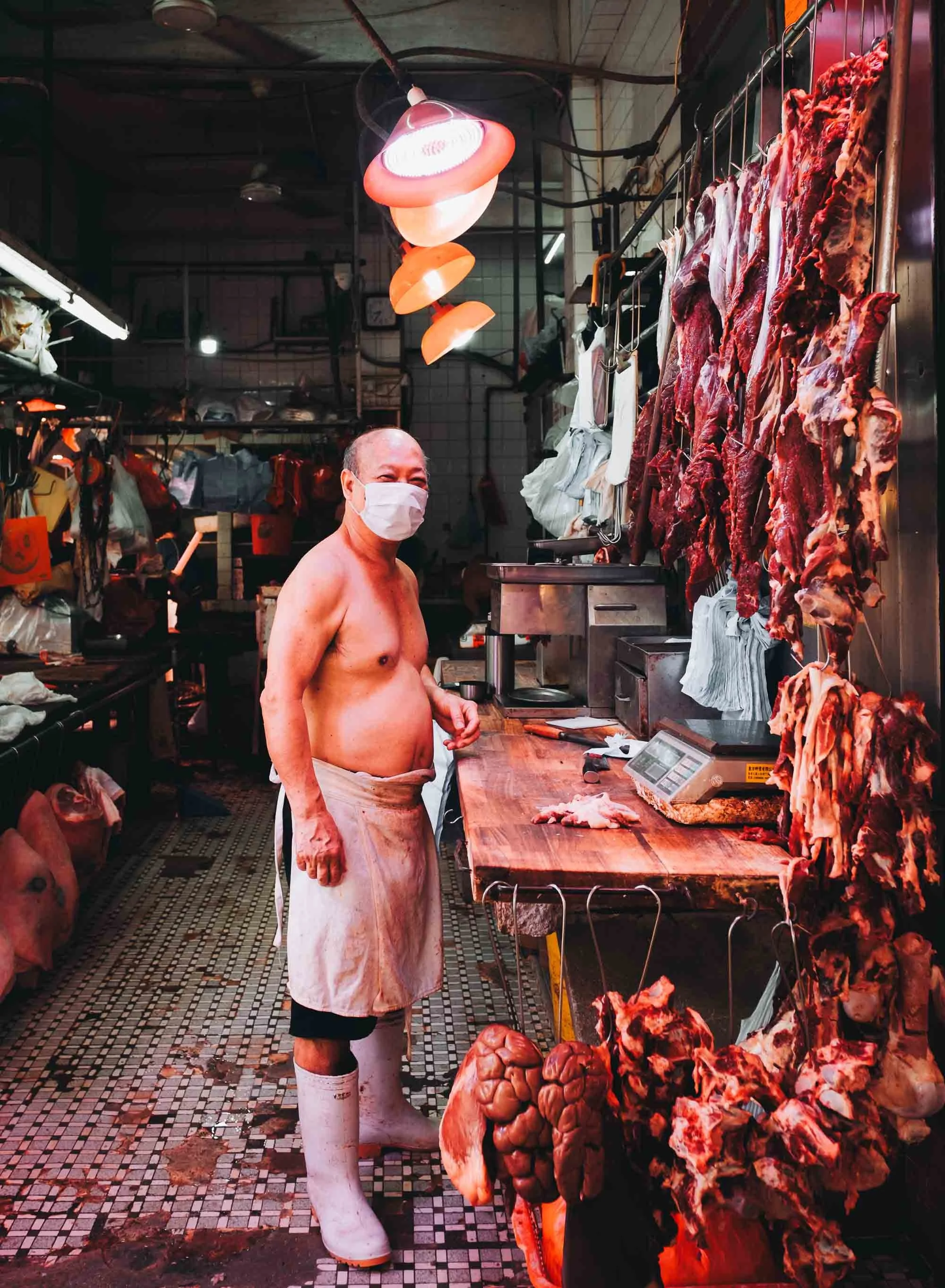A shirtless man wearing a face mask and apron standing in a Hong Kong butcher shop surrounded by hanging and sliced raw meat.