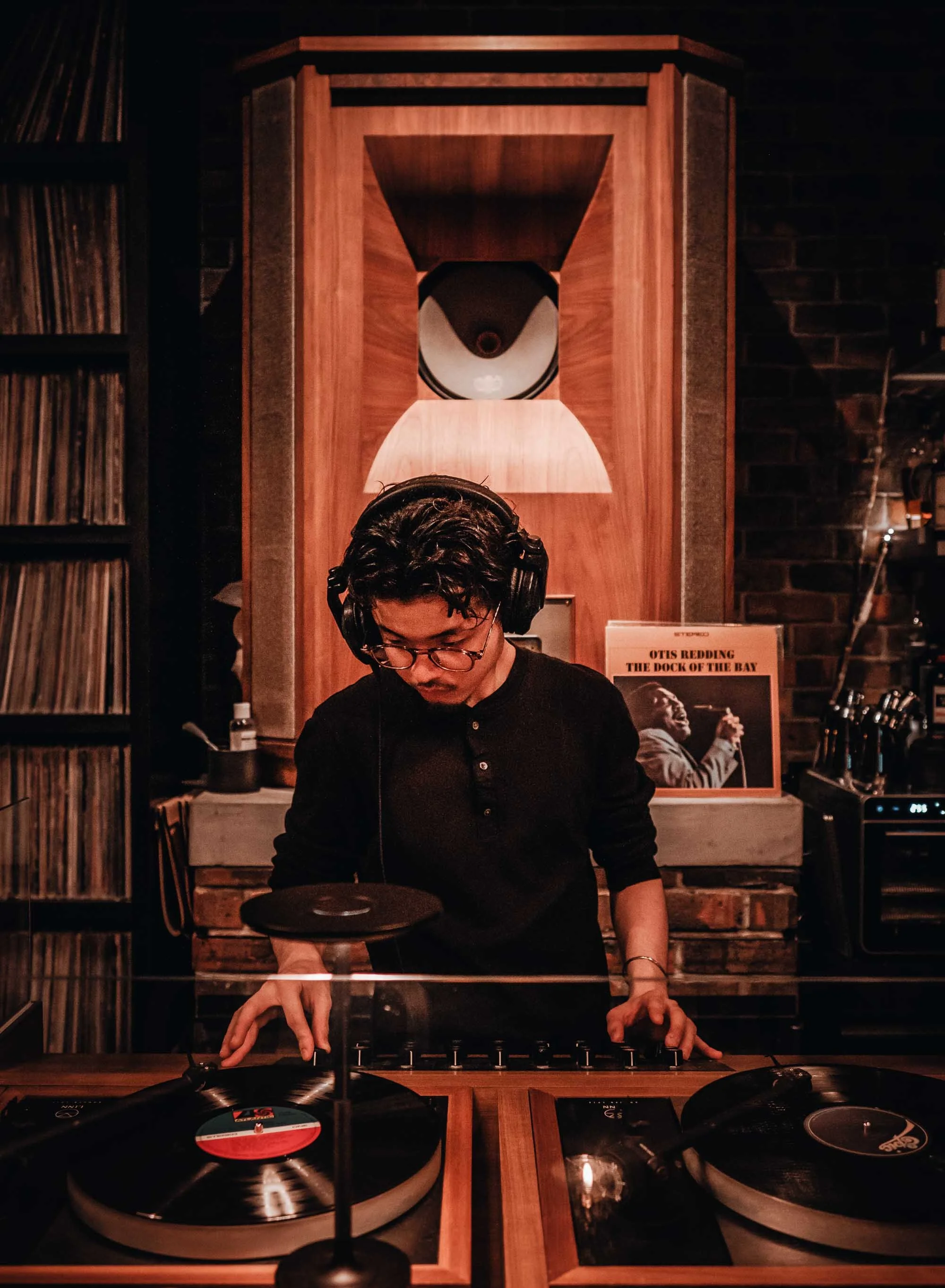 A DJ wearing headphones and glasses is mixing records at a turntable in a dimly lit room with wooden and brick decor, including a vinyl record album cover on display.