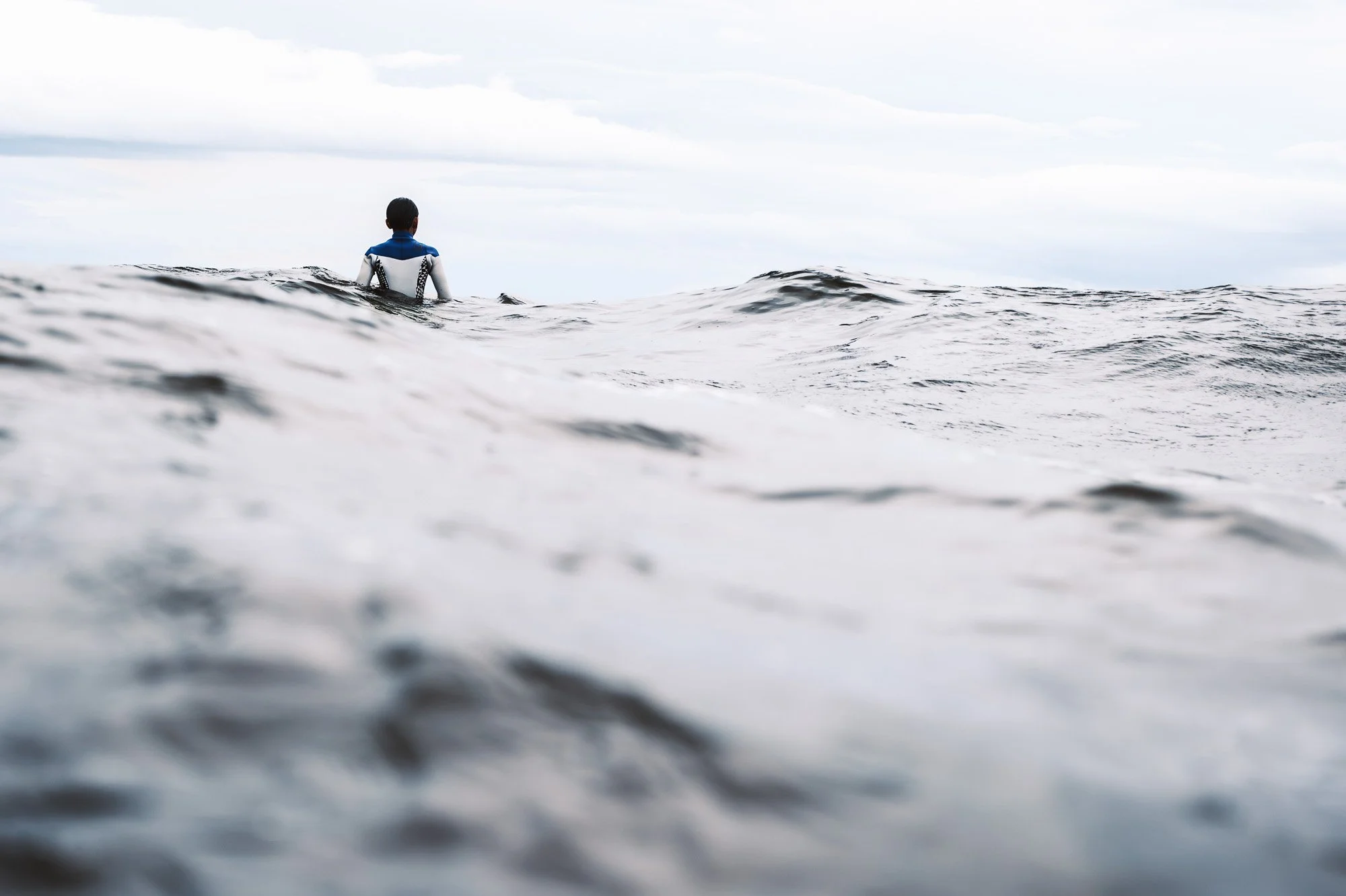 A person in a wetsuit sitting on a surfboard in the ocean, gazing at the cloudy sky in the distance.