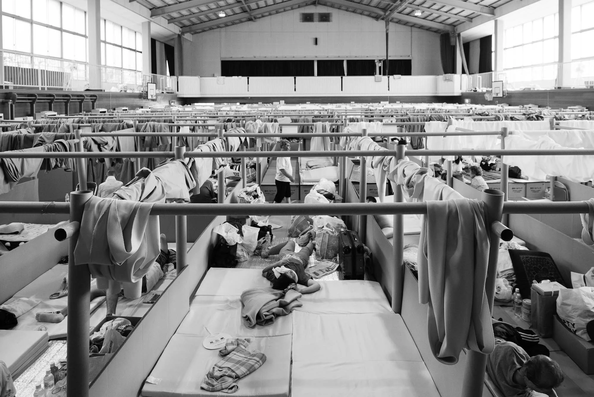 Black and white photo of a crowded indoor shelter with many beds, showing some people lying down, resting or sleeping, surrounded by personal belongings.