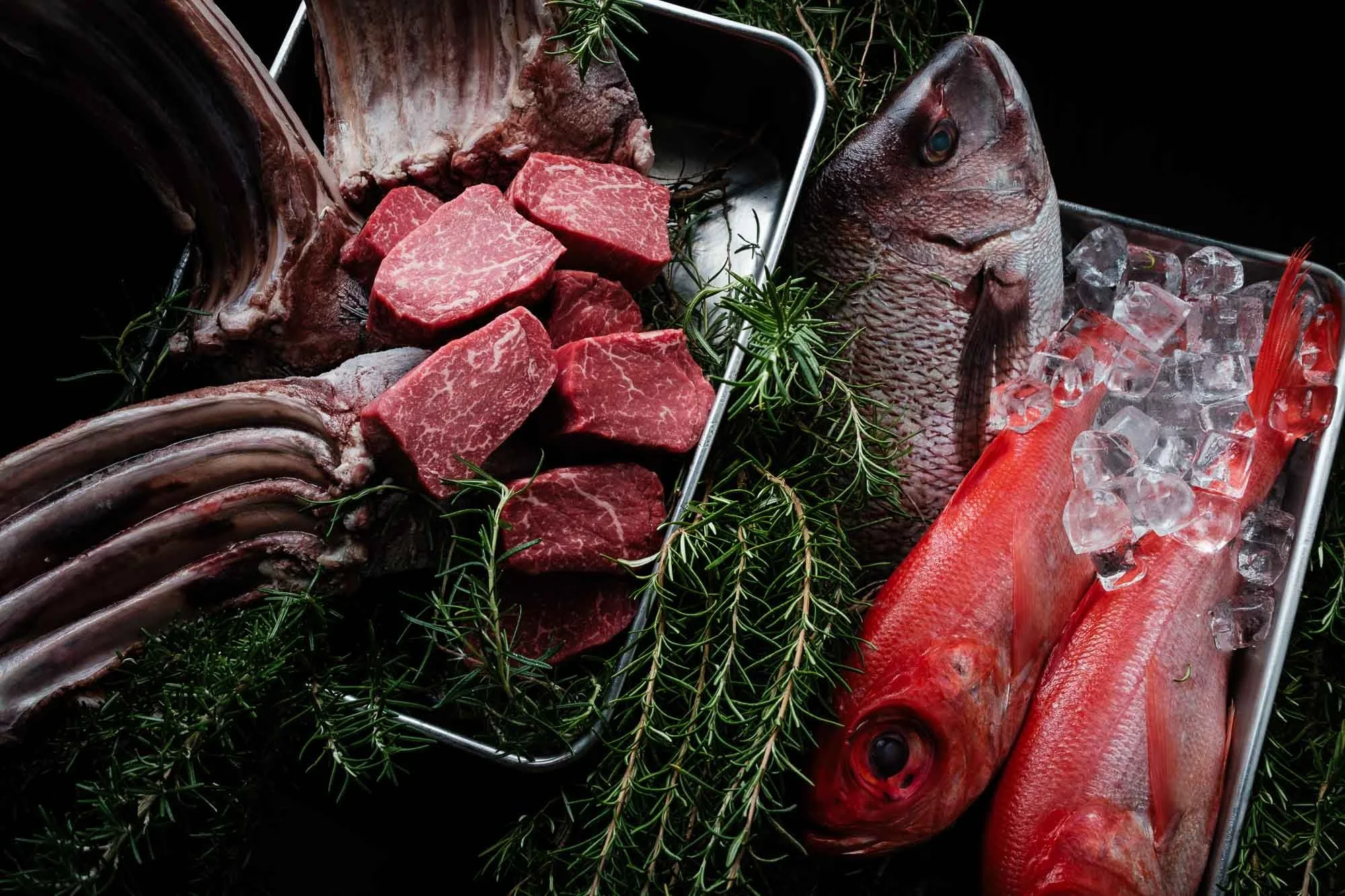 Fresh cuts of red meat, whole fish on ice, and sprigs of rosemary in a display tray.